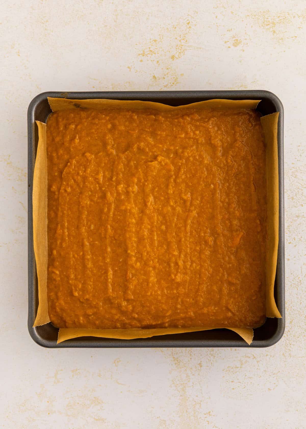 A square baking pan holds sweet potato pie bars batter, lined with parchment paper.