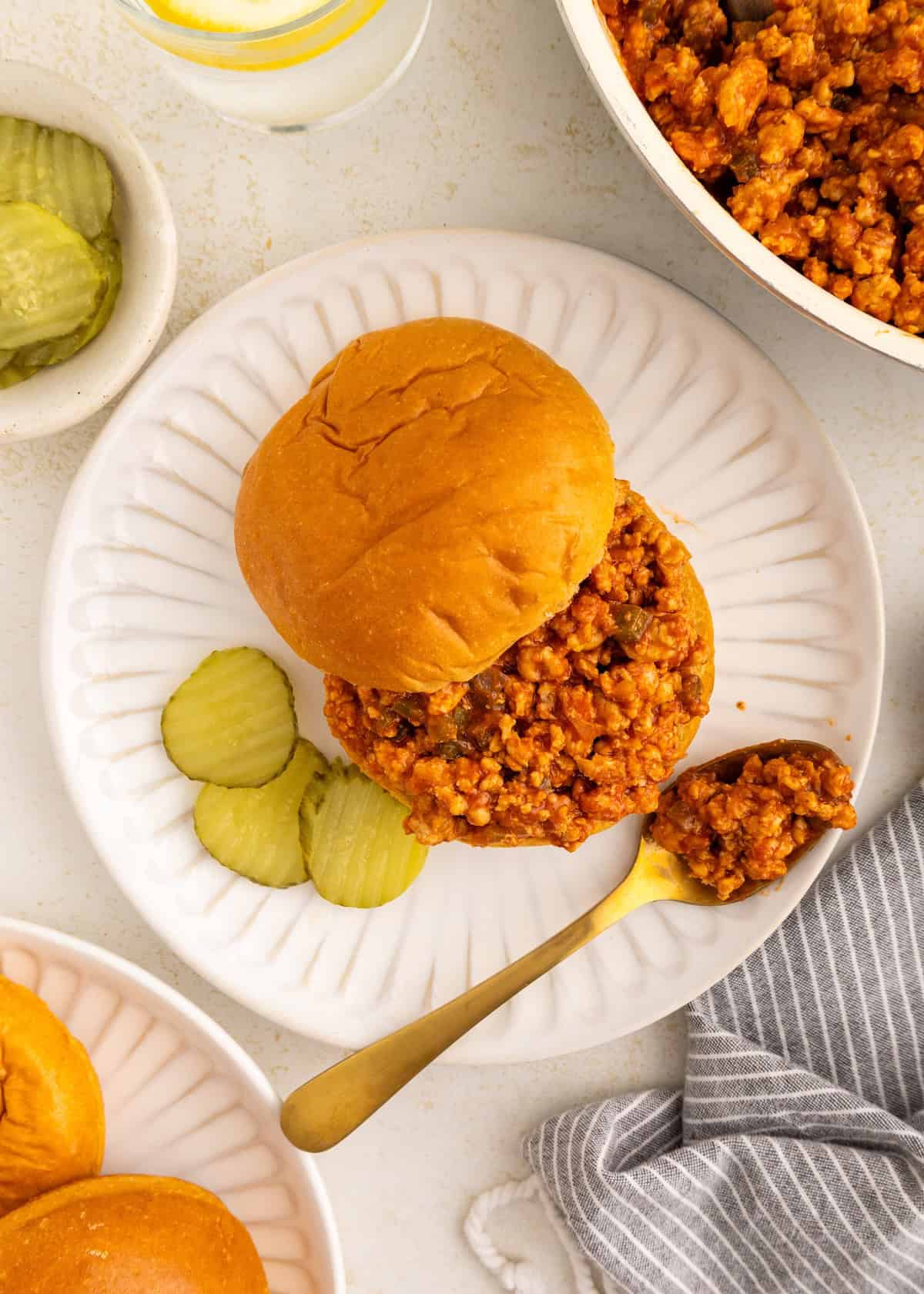 Turkey sloppy joe sandwich on a plate with pickle slices and a gold spoon, overhead view.