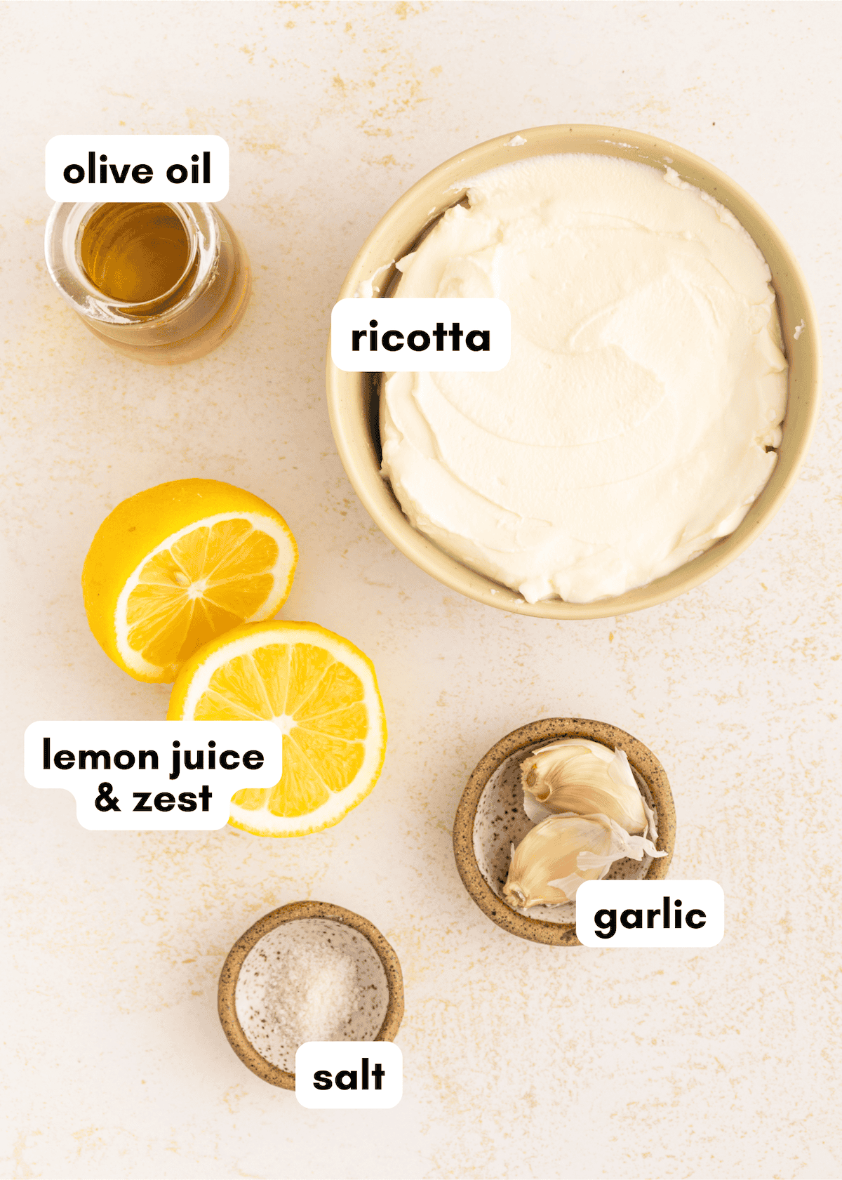 Bowl of whipped ricotta dip with olive oil, lemon, garlic, and salt on a light surface with labels.