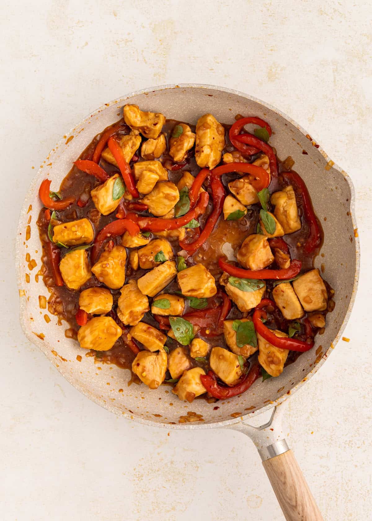 Thai Basil Chicken with red bell peppers and basil cooking in a skillet with sauce on a light background.