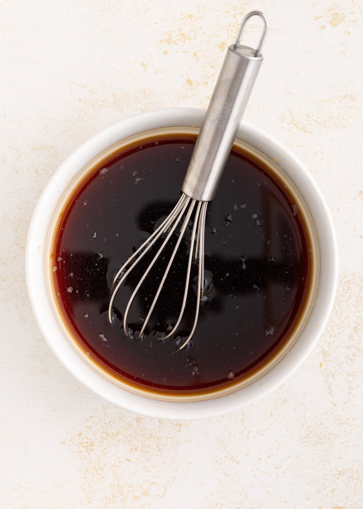 A metal whisk in a white bowl filled with dark brown liquid sauce.