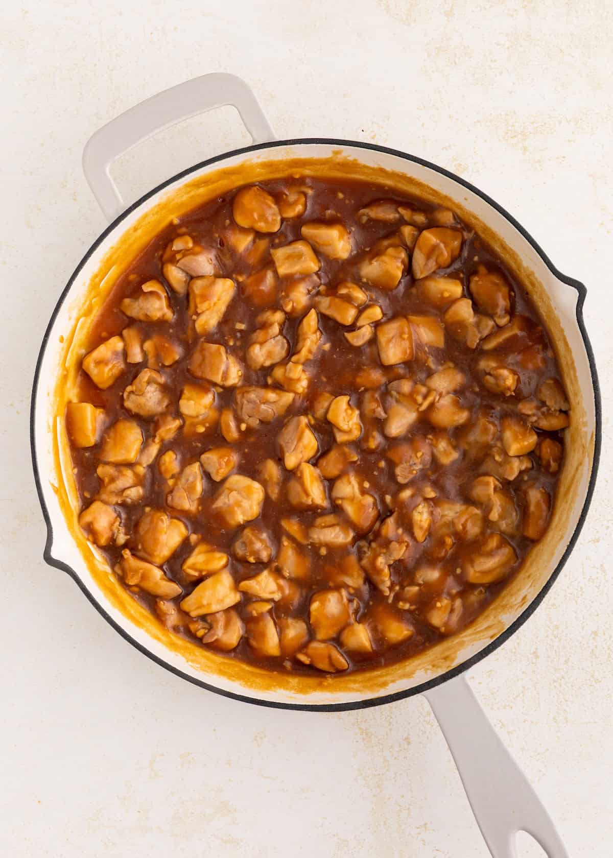 Chicken pieces in a thick brown sauce cooking in a white pot on a light surface.