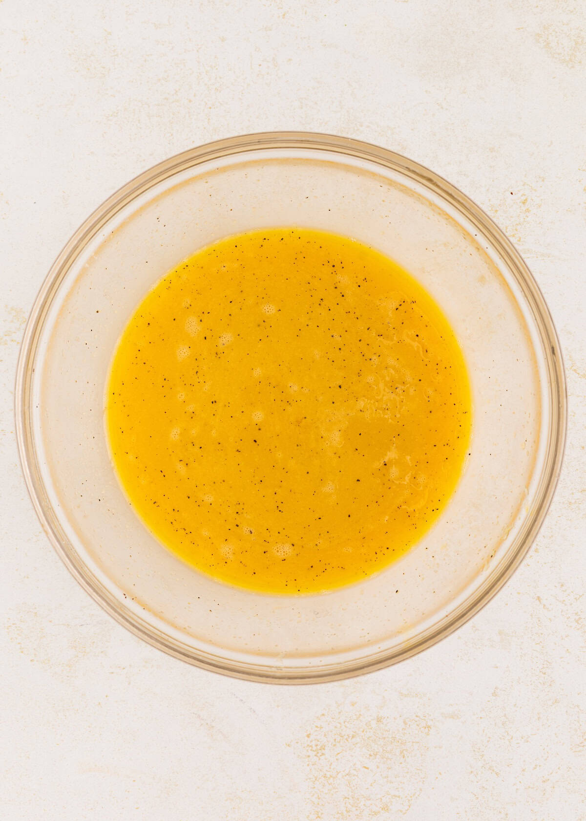 Clear bowl with whisked eggs and black pepper on a white surface.