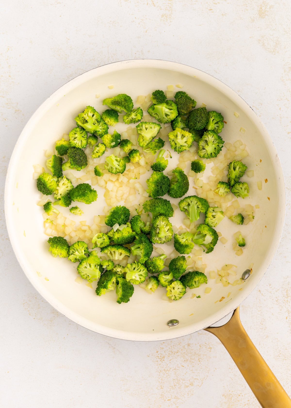 Broccoli and chopped onions cooking in a white skillet with a gold handle.