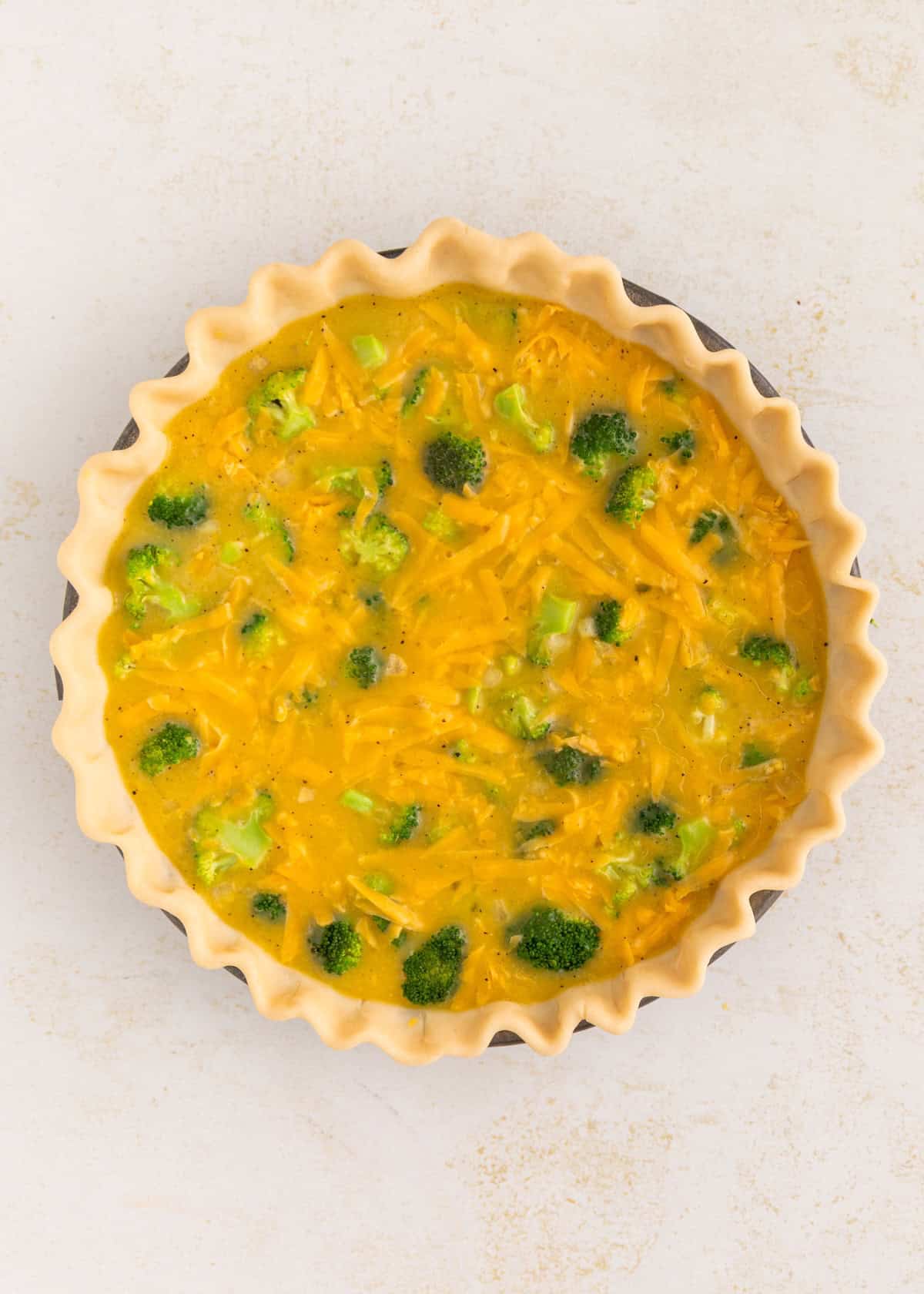 Unbaked quiche with broccoli and cheese in a pie crust on a light surface.