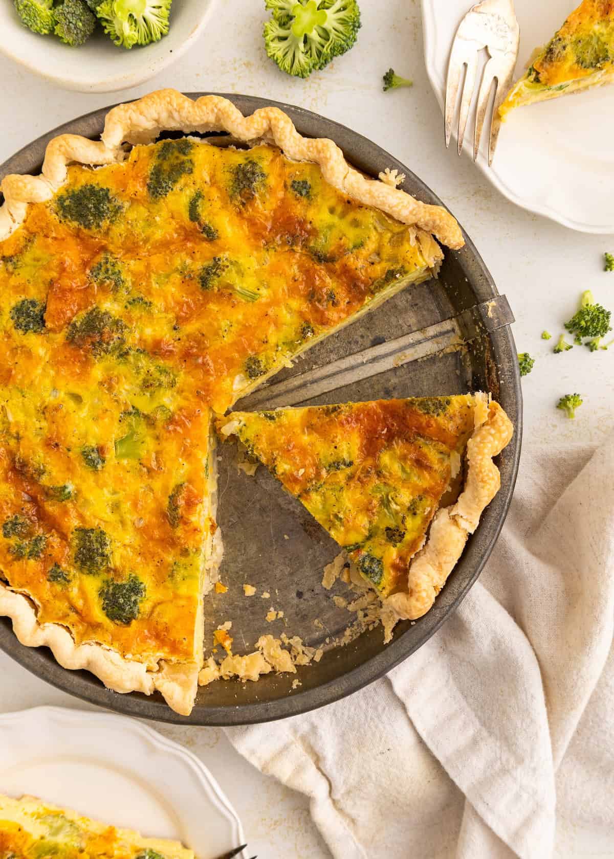 Broccoli quiche with a slice missing, in a baking pan on a light table with plates nearby.