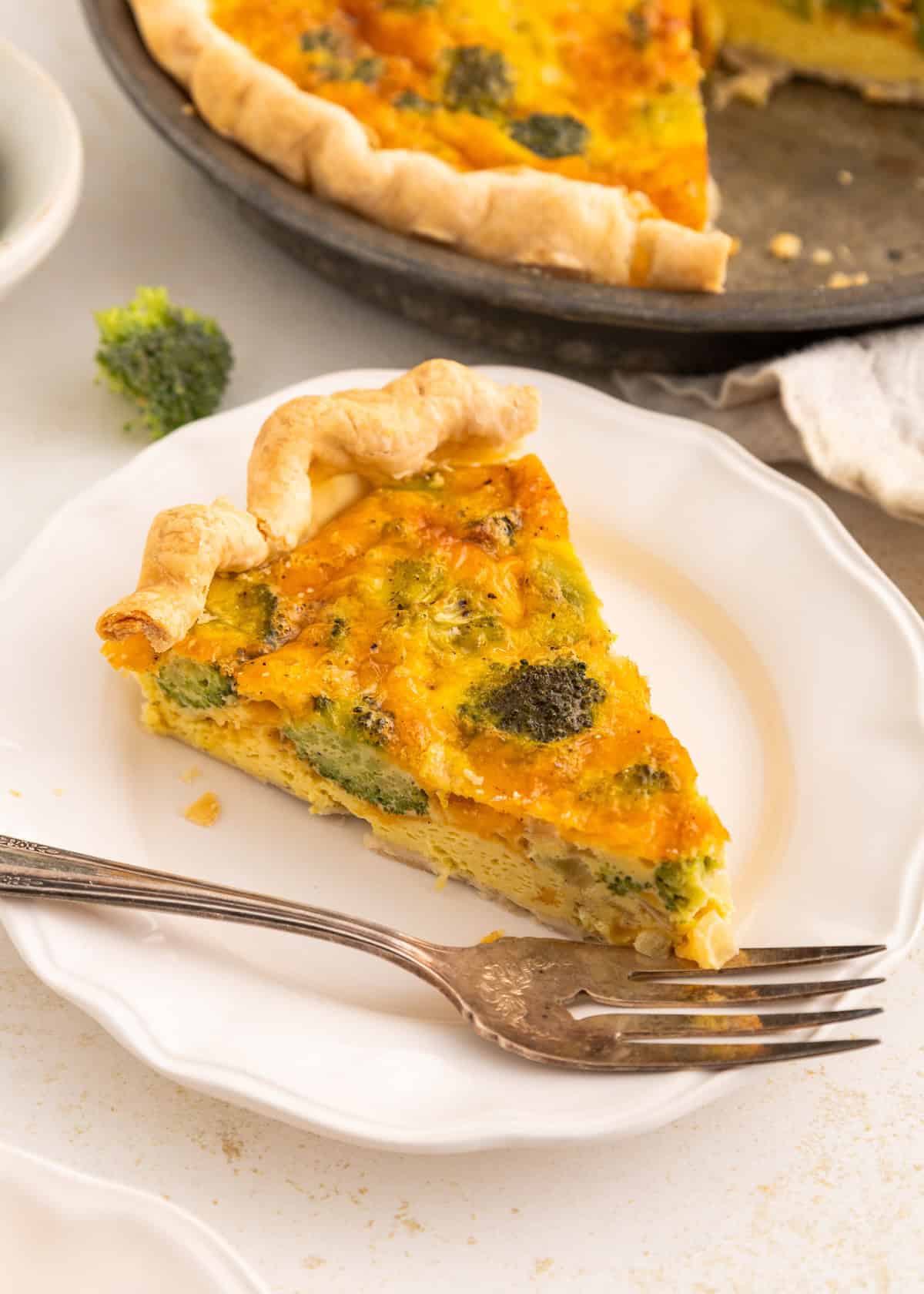 A slice of broccoli quiche on a white plate with a fork beside it.