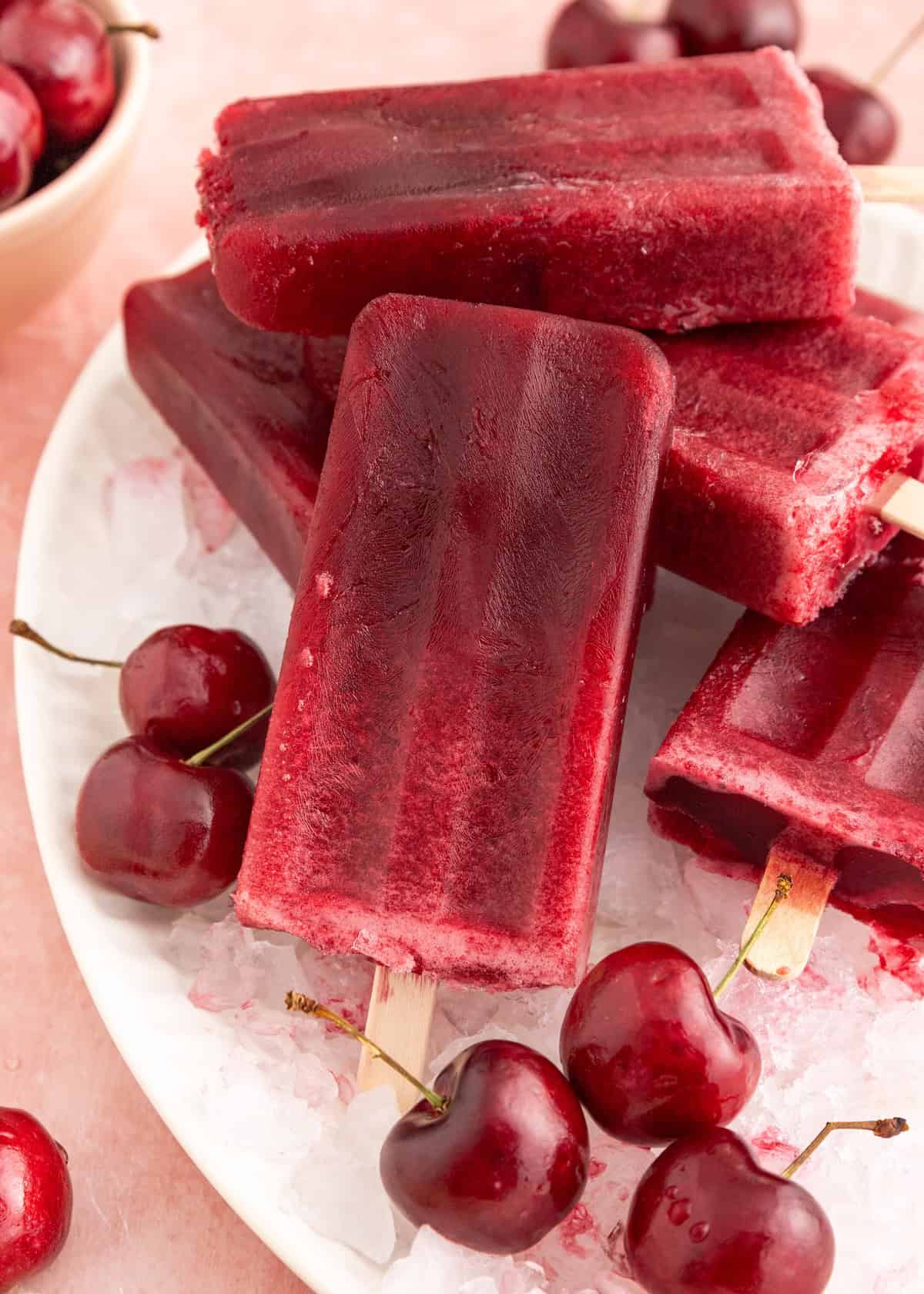 Cherry popsicles stacked on ice with fresh cherries scattered around on a white plate.