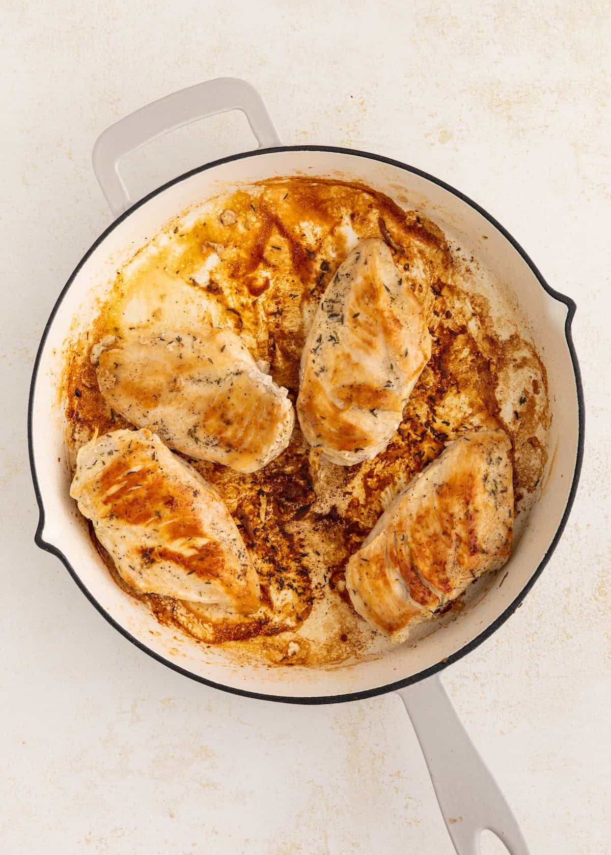Four cooked chicken breasts in a white skillet with browned seasoning and juices.