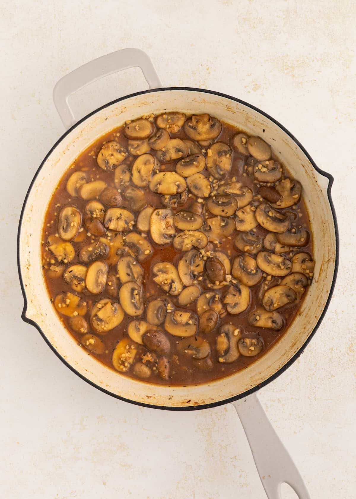 Sliced mushrooms simmering in a brown sauce inside a large white pot on a light surface.