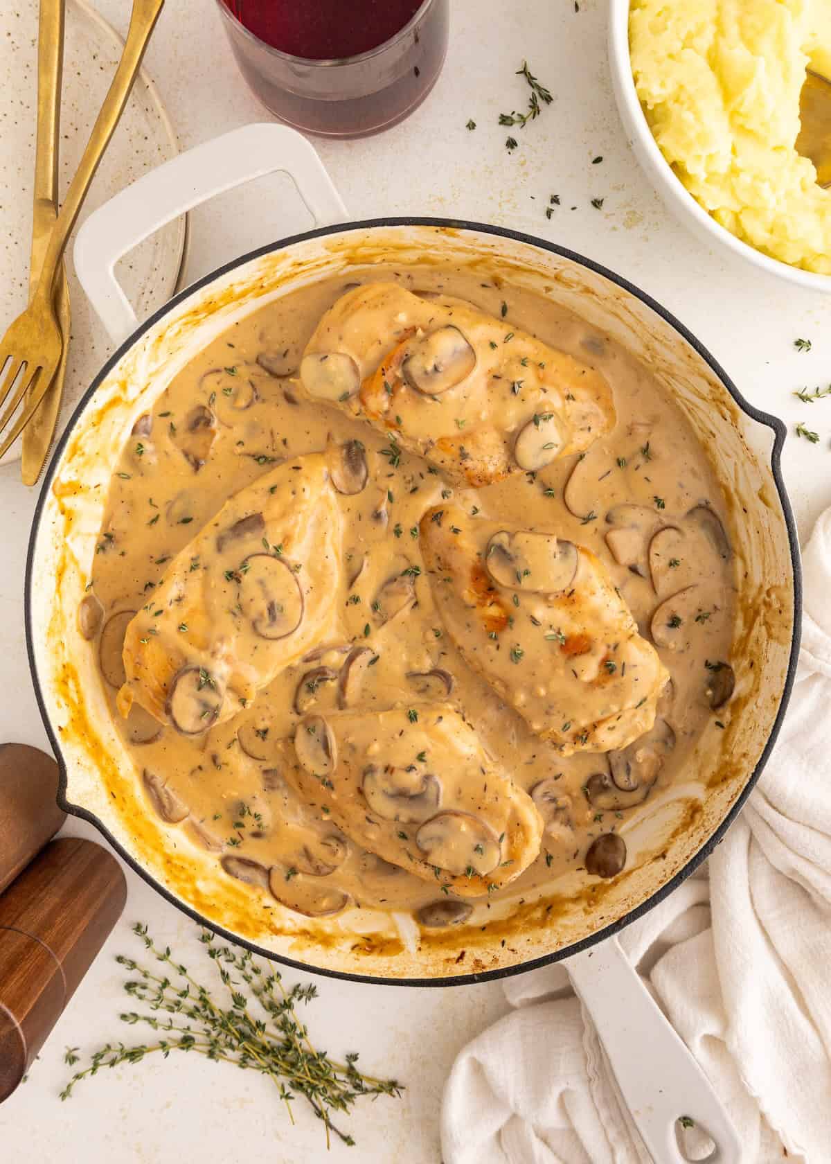 Chicken breasts in creamy mushroom sauce in a pan, with mashed potatoes and herbs on the side.