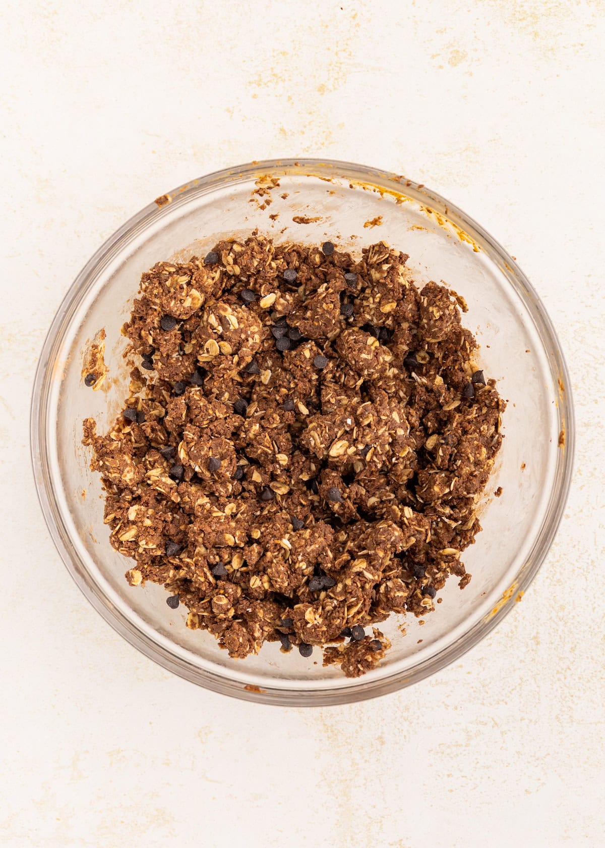A glass bowl filled with chunky chocolate oat cookie dough on a light surface.