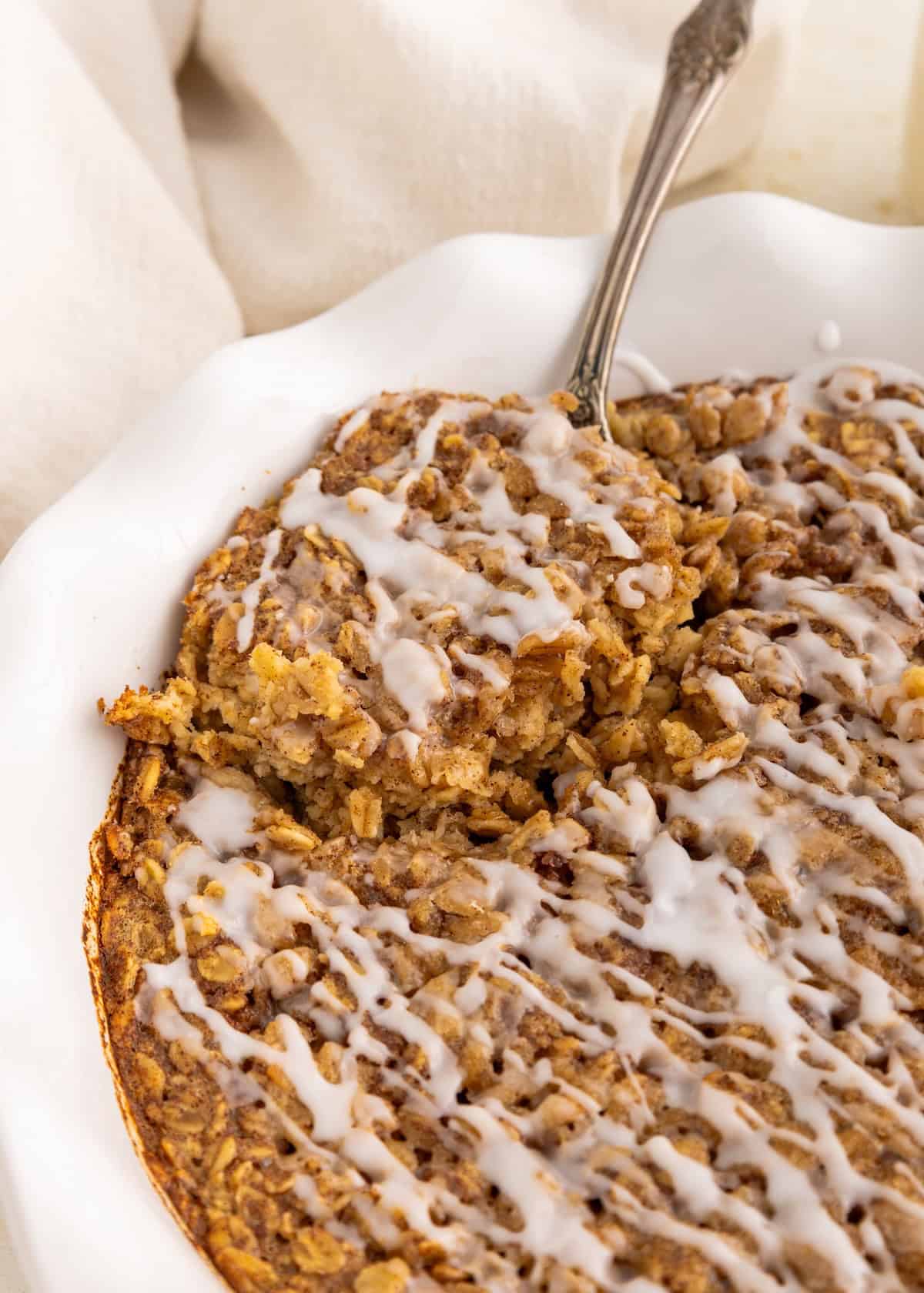 Cinnamon Roll Baked Oatmeal drizzled with icing in a white dish, spoon taking a portion.