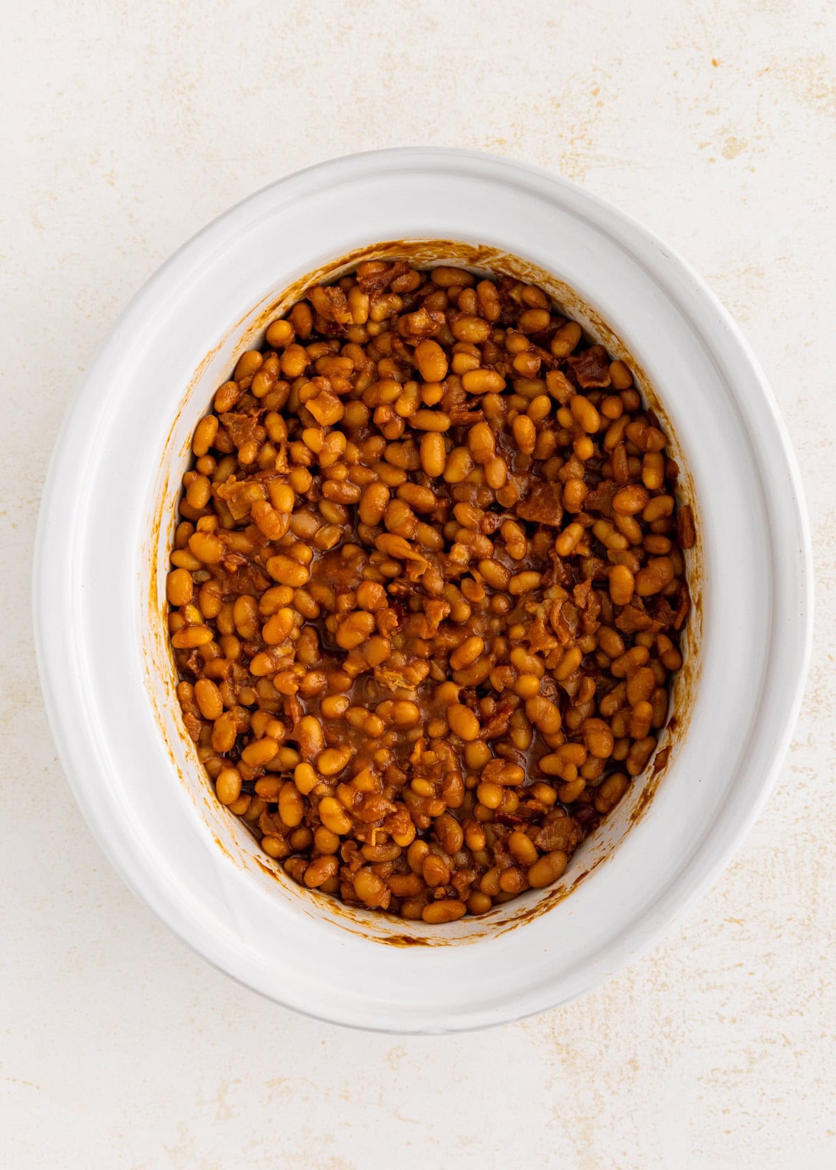 Baked beans in a white oval dish on a light-colored surface, viewed from above.