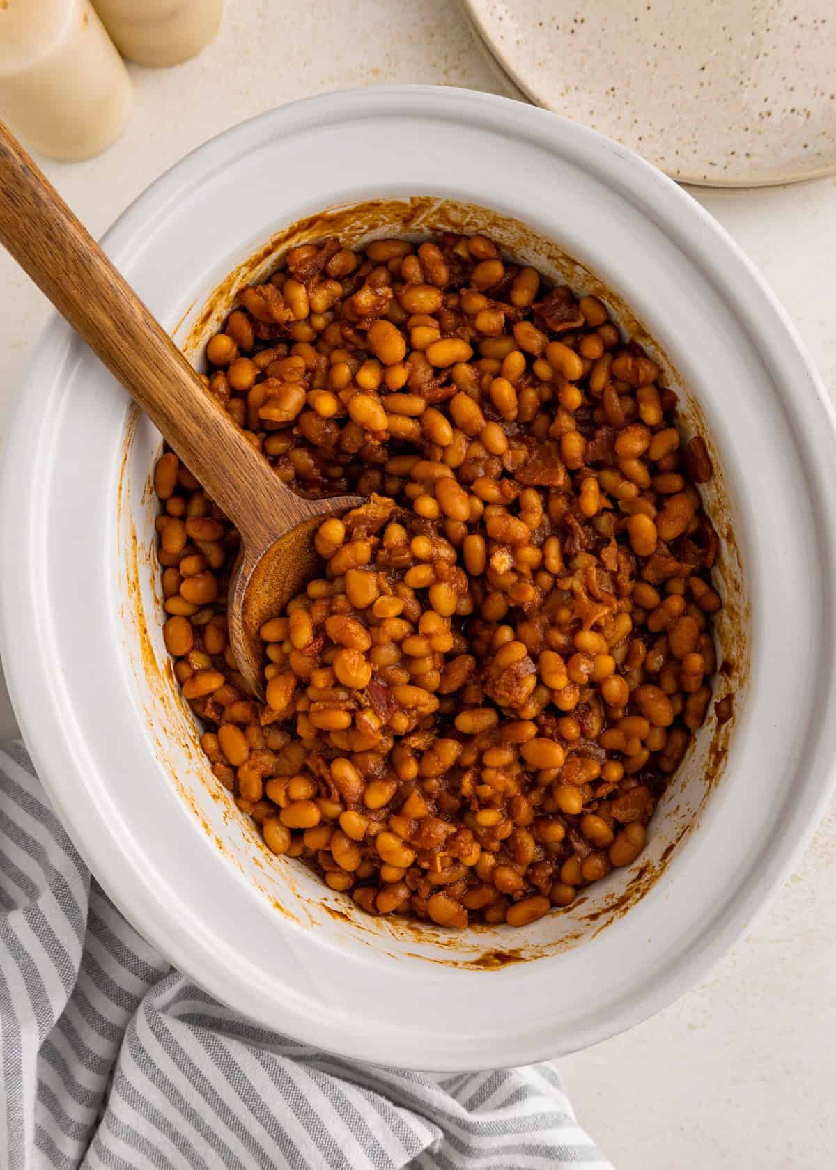 Baked beans in a white dish with a wooden spoon, next to a striped towel.