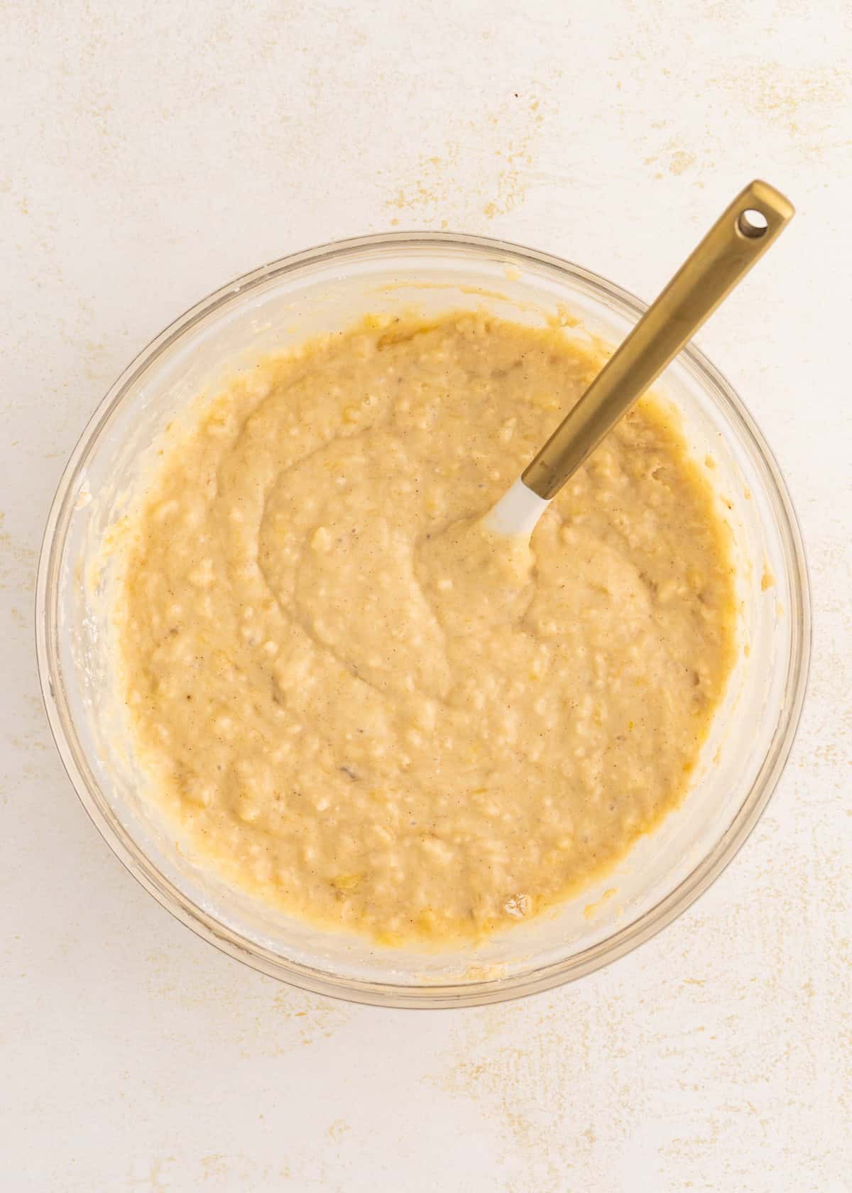 Overhead view of banana bread batter in a glass bowl with a golden spatula.