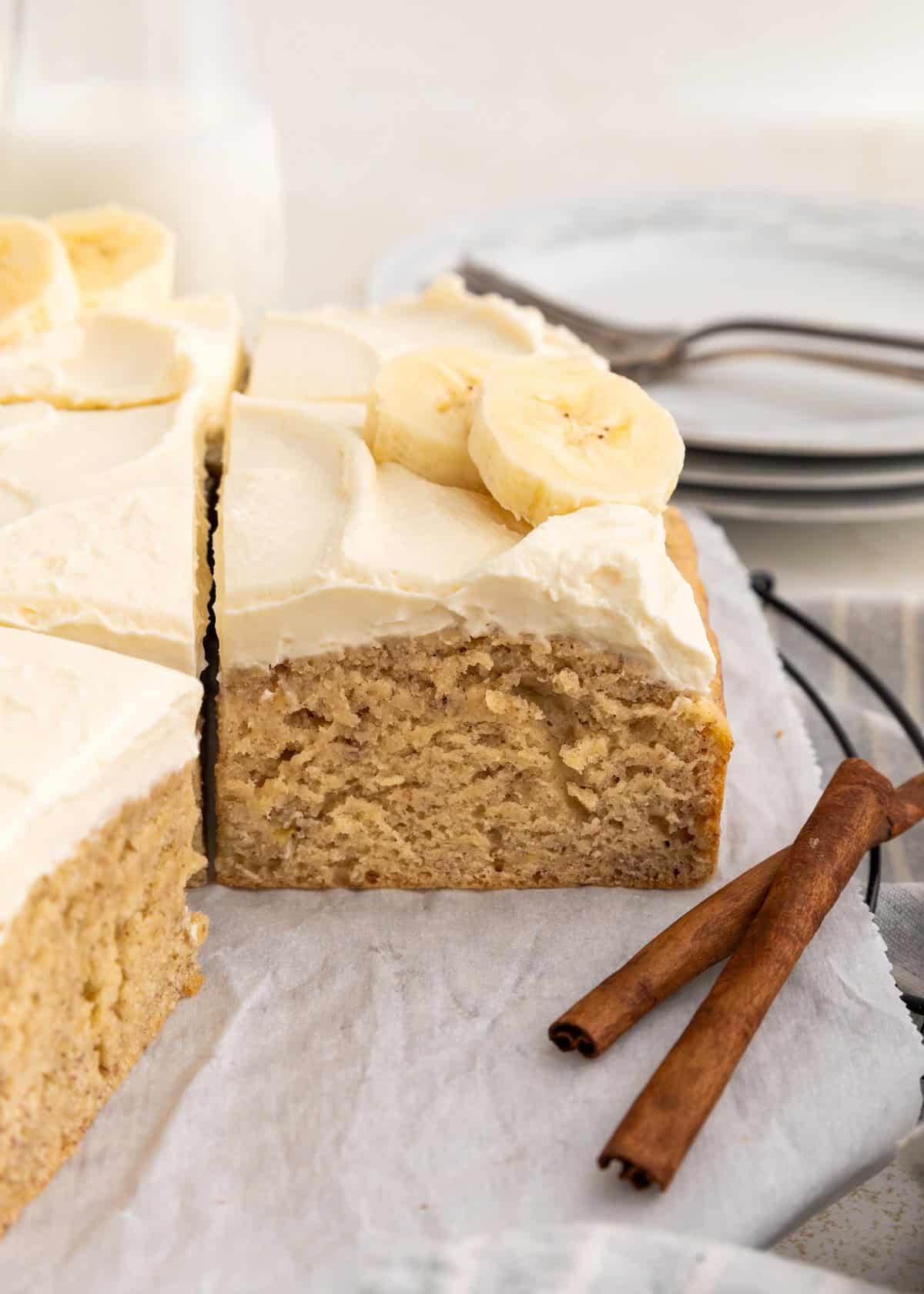 Sliced banana cake with cream cheese frosting, banana slices, and cinnamon sticks.