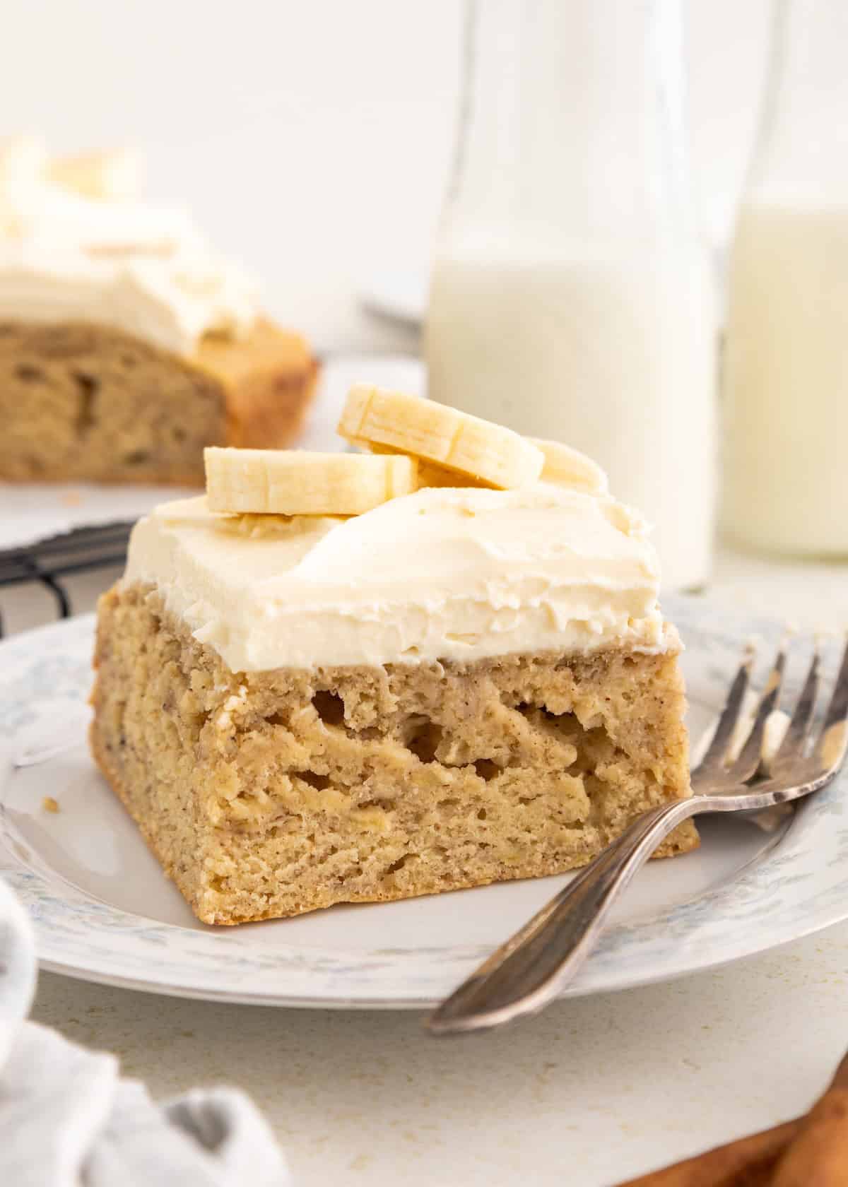 A slice of banana cake with cream cheese frosting and fresh banana slices on a plate with a fork.