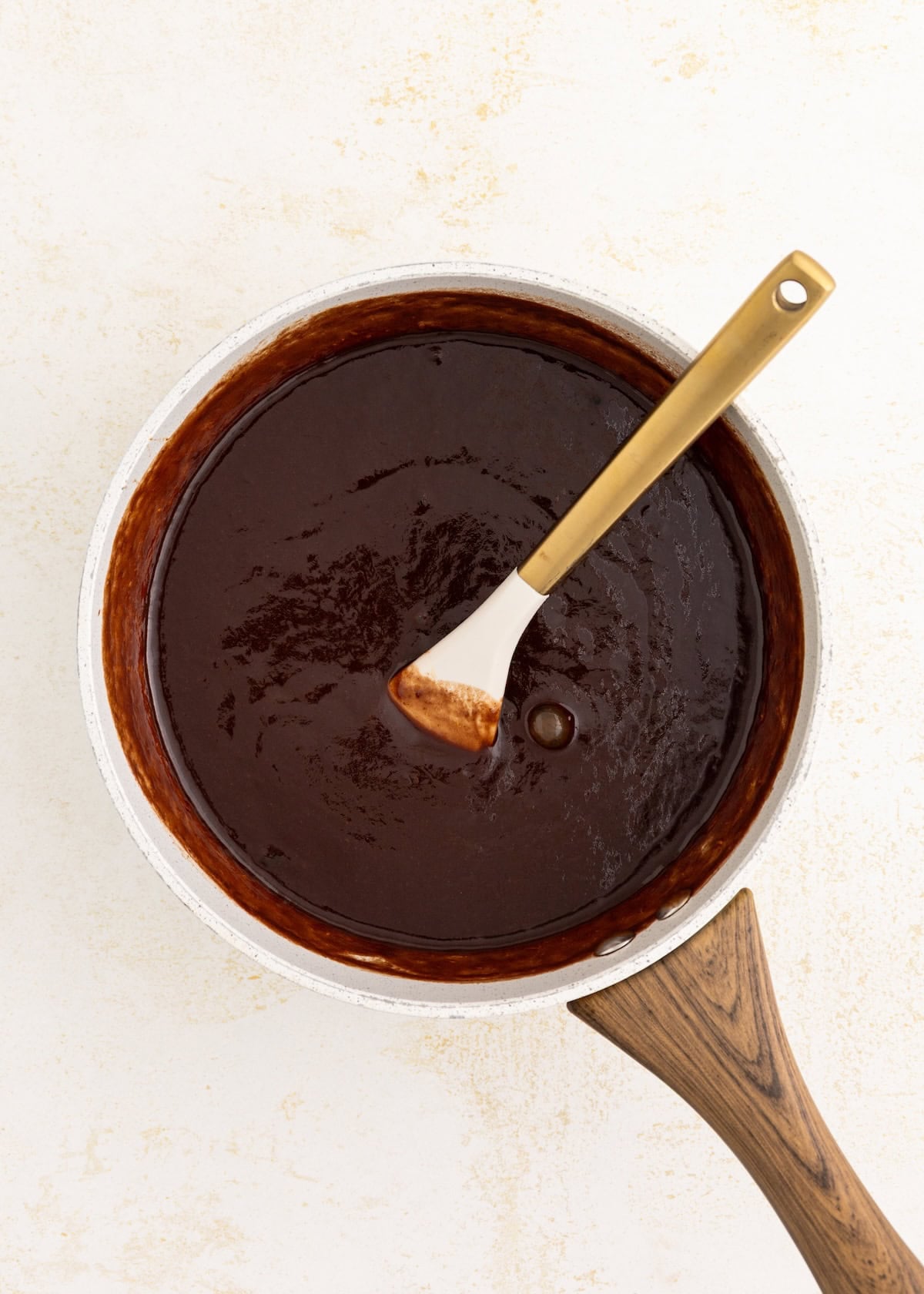 A saucepan of chocolate sauce with a spatula on a light surface.