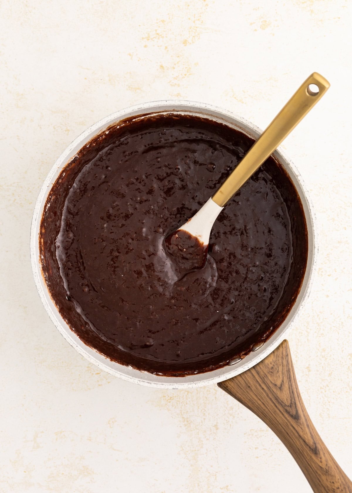 A saucepan filled with chocolate brownie batter and a spatula on a light surface.