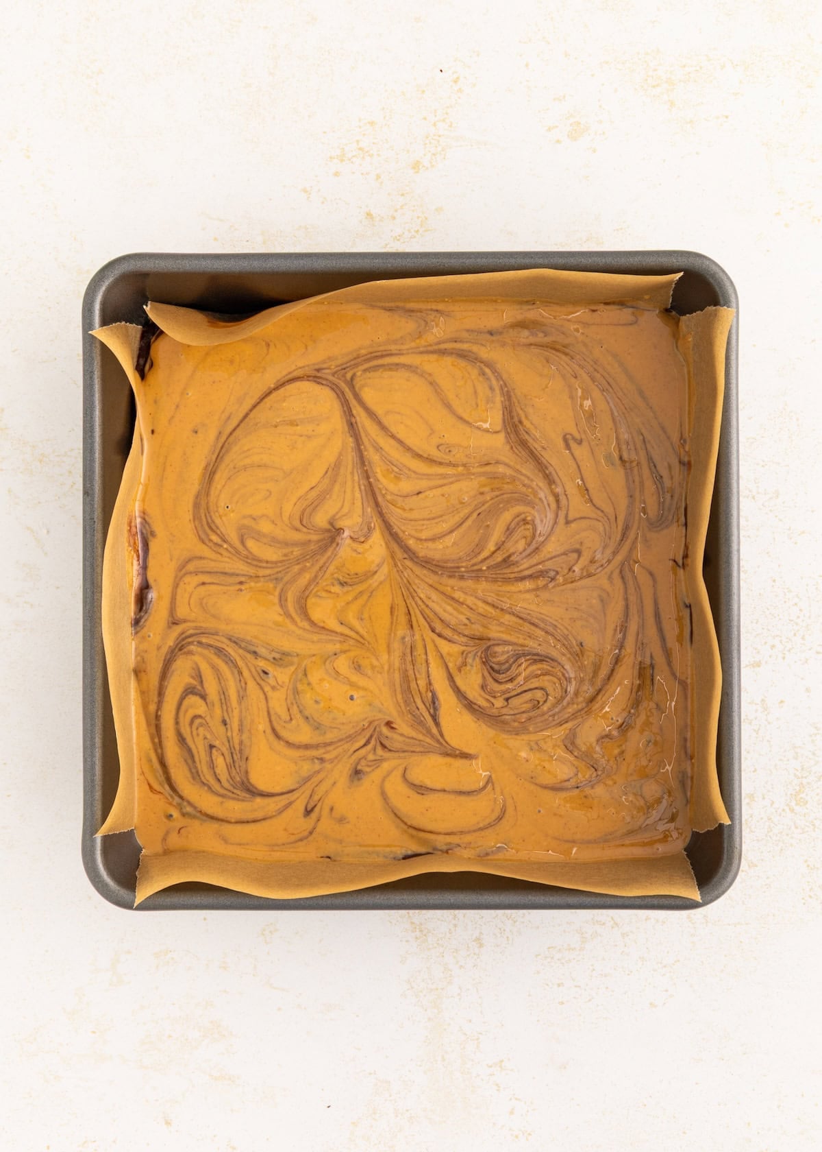 Square pan of swirled brown batter on parchment paper, viewed from above.