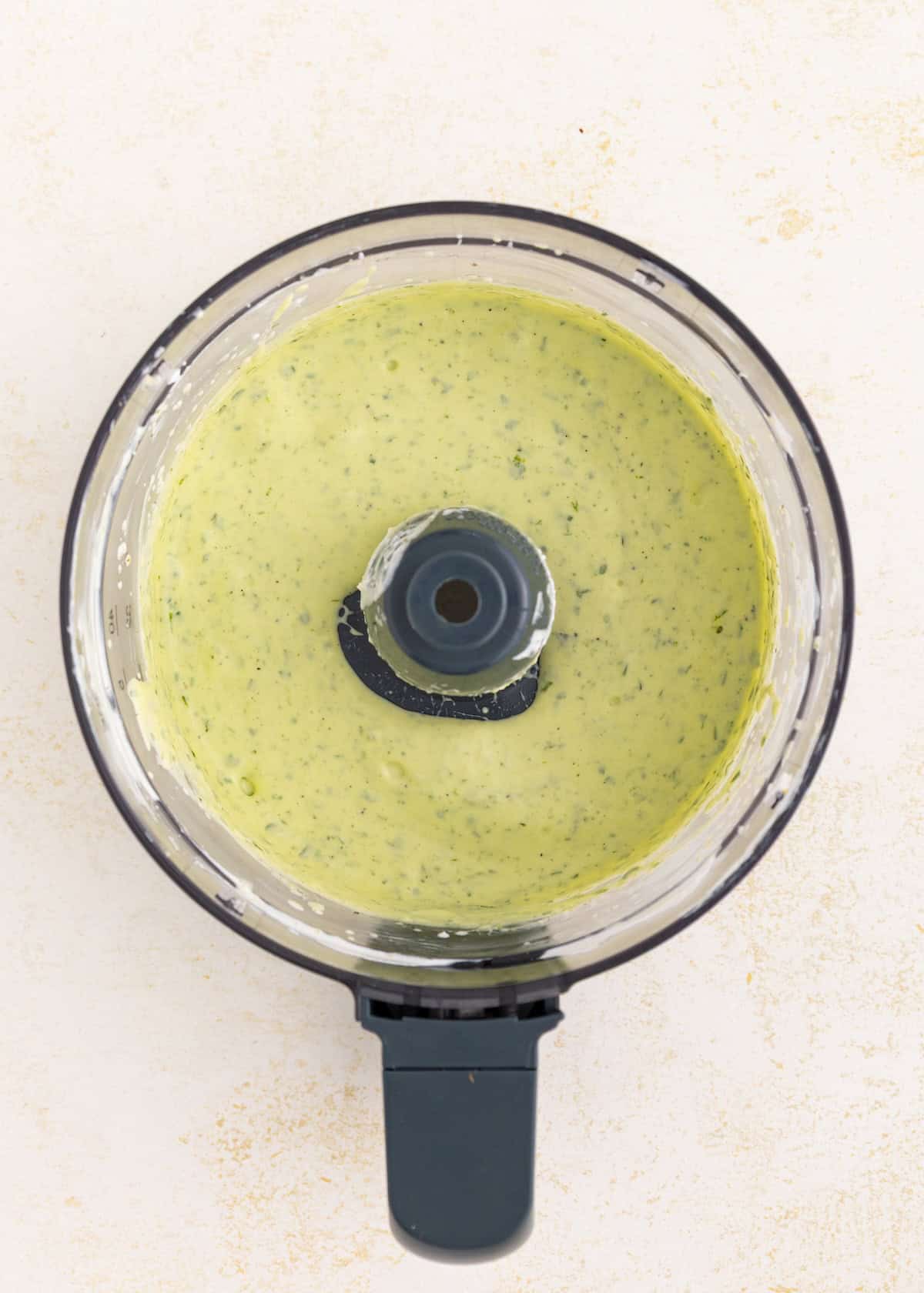 Overhead view of creamy green sauce in a food processor on a light background.