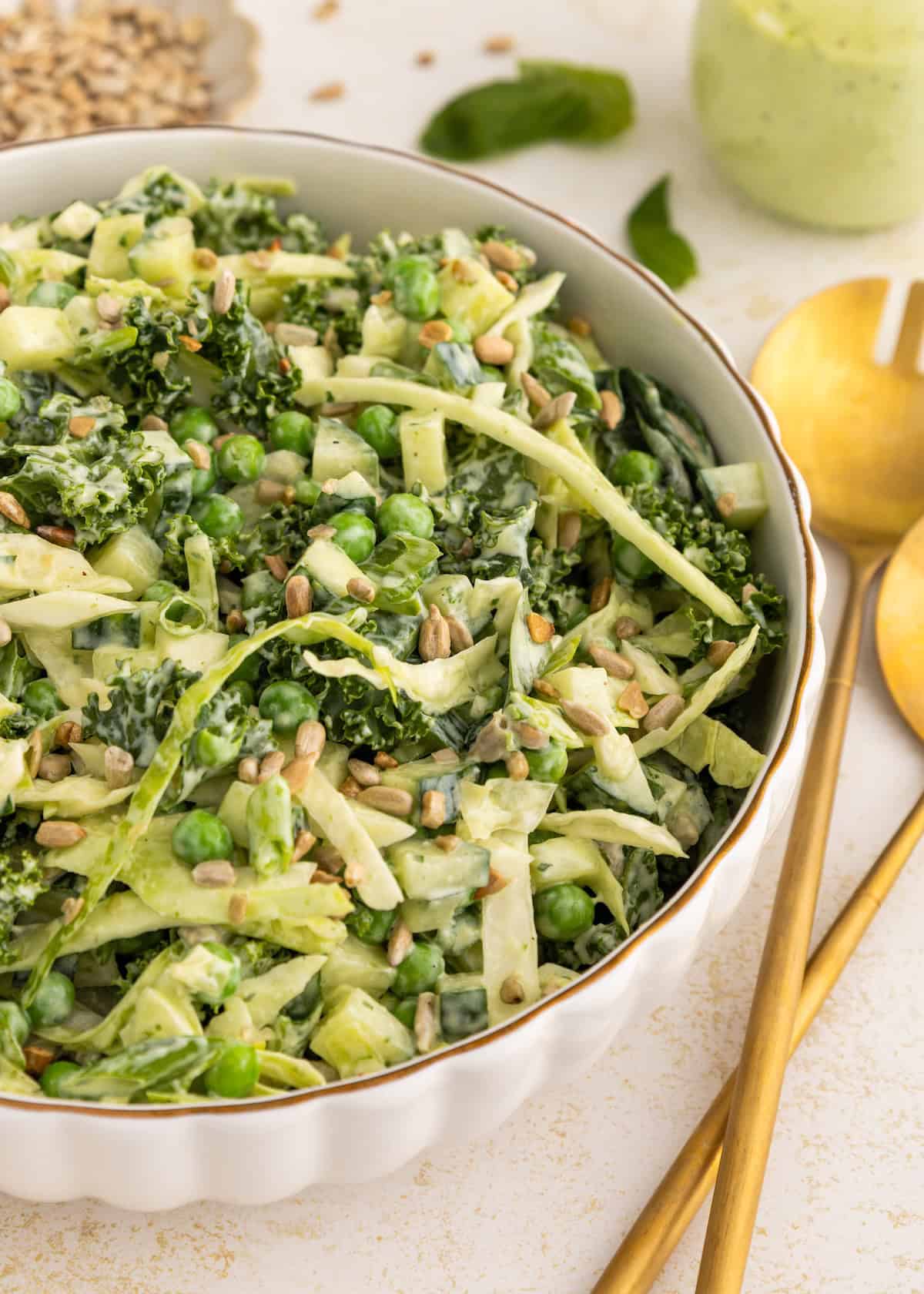 A bowl of creamy green salad with kale, peas, sunflower seeds, and golden utensils beside it.