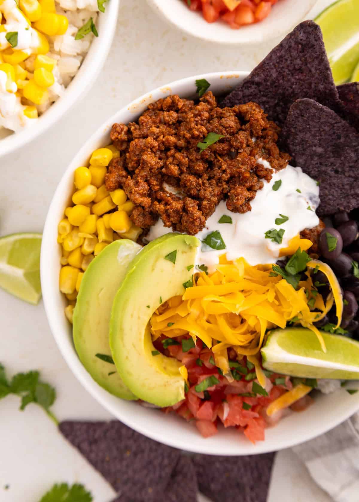 A taco bowl with ground meat, corn, avocado, cheese, black beans, and lime wedges.