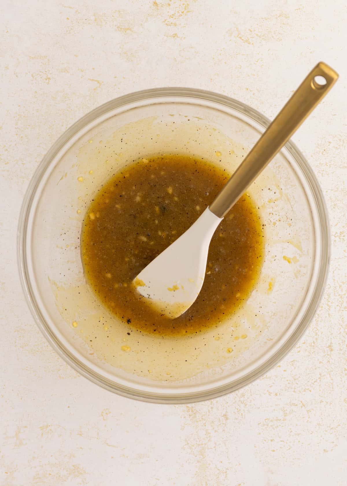 Clear bowl with brown vinaigrette and a white spatula on a light surface.