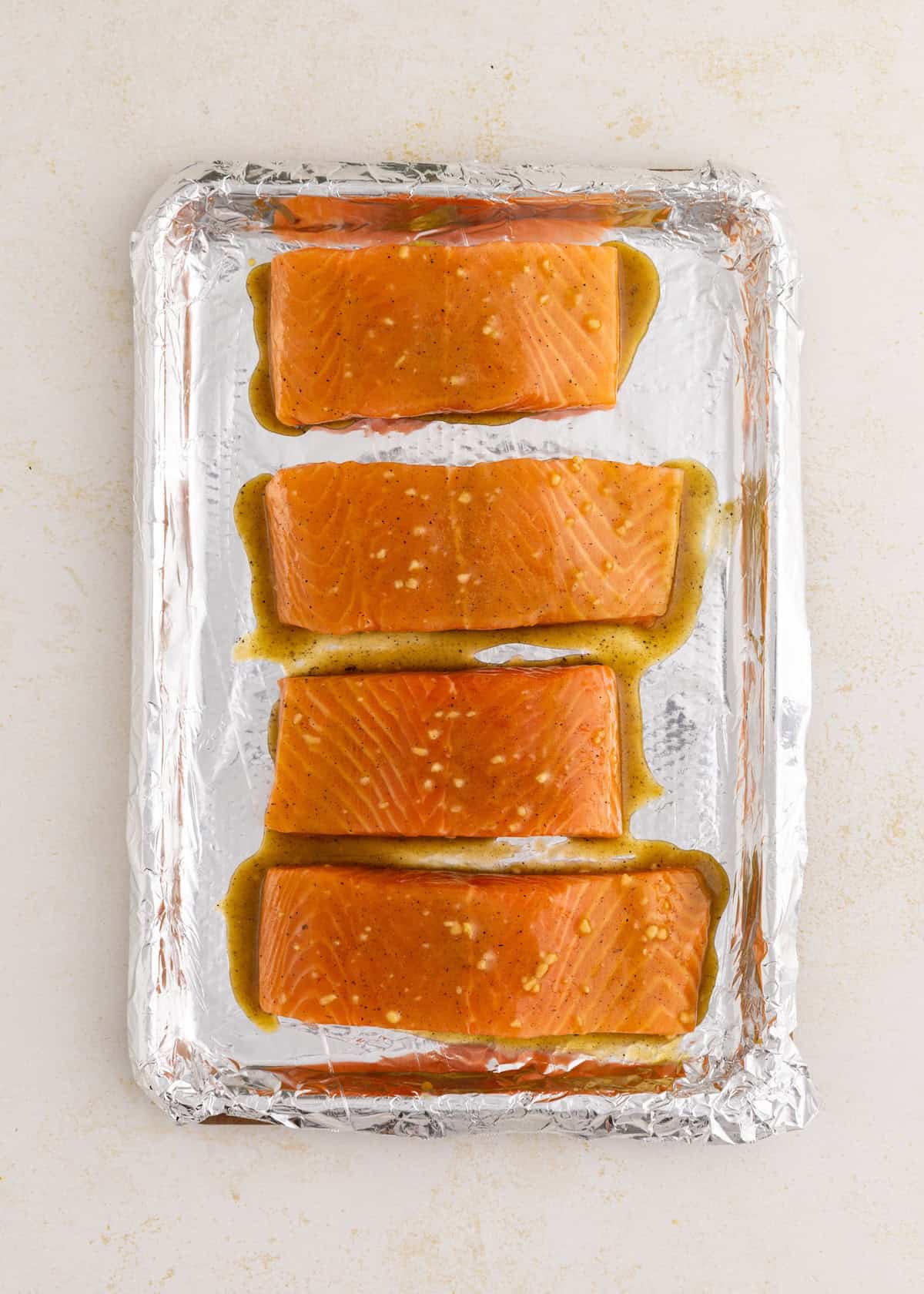 Four raw salmon fillets with marinade on a foil-lined baking sheet.