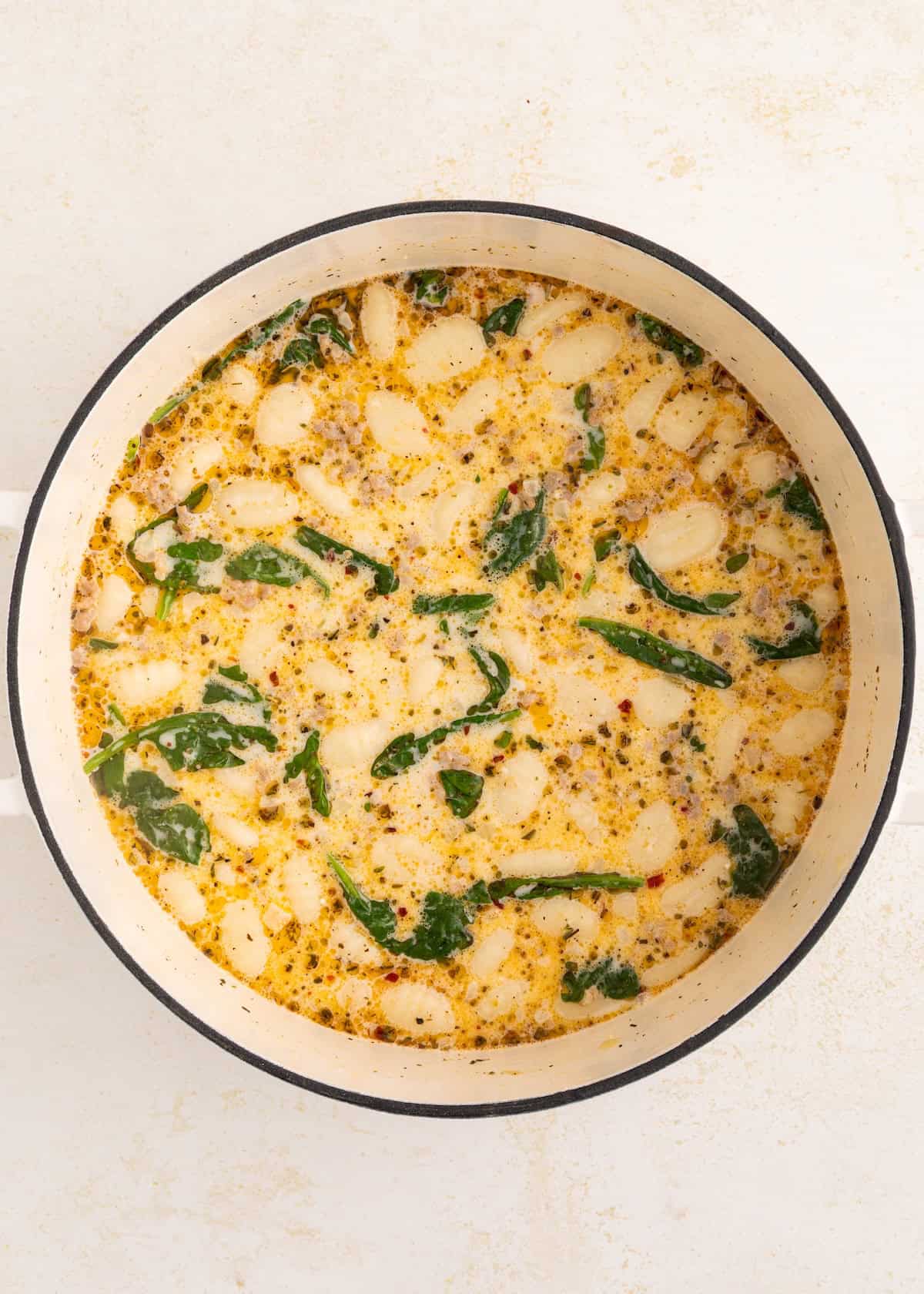 Creamy soup with gnocchi, spinach, and herbs simmering in a white pot on a light surface.