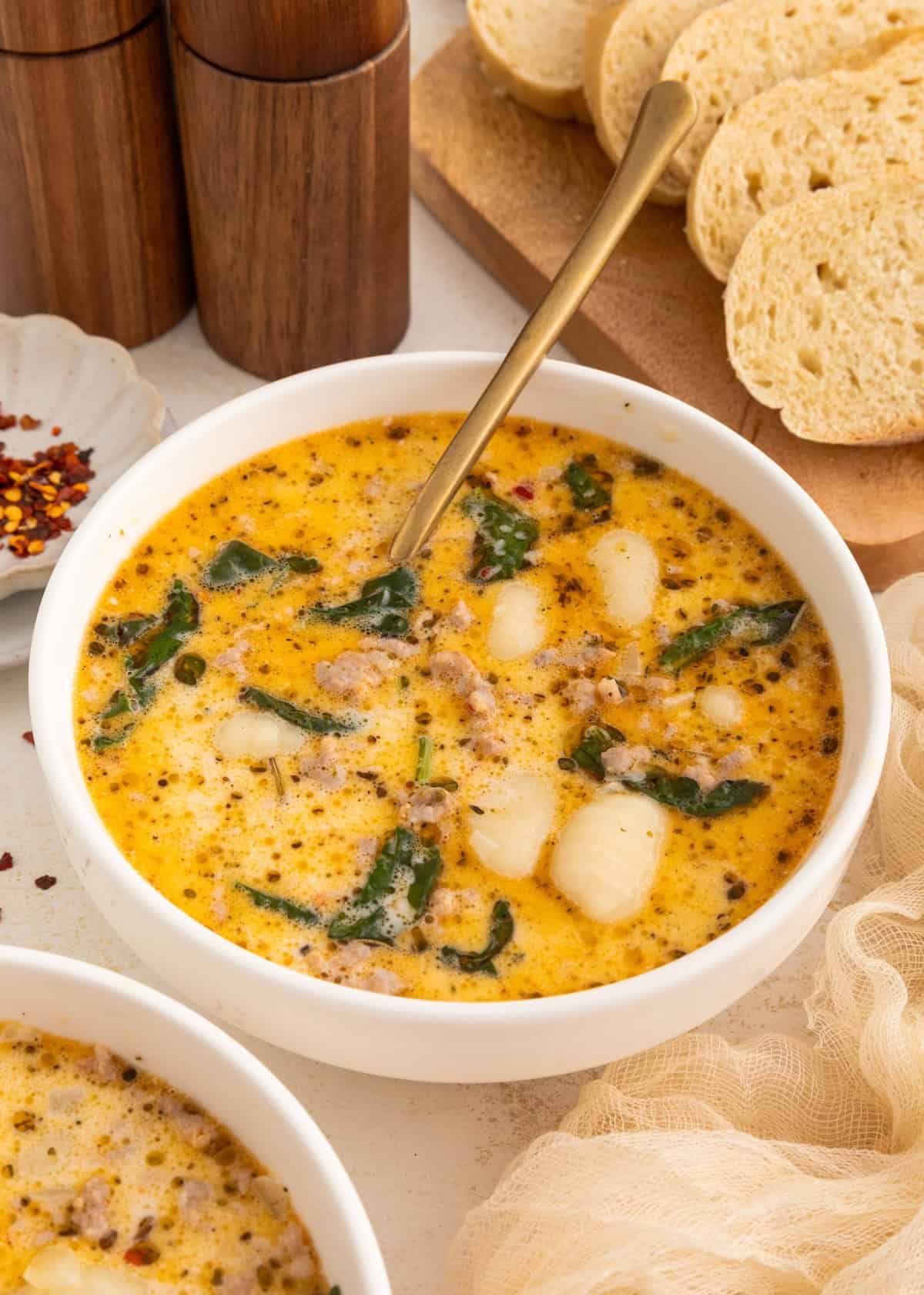 Creamy soup with sausage, gnocchi, and spinach in a white bowl, bread slices in background.