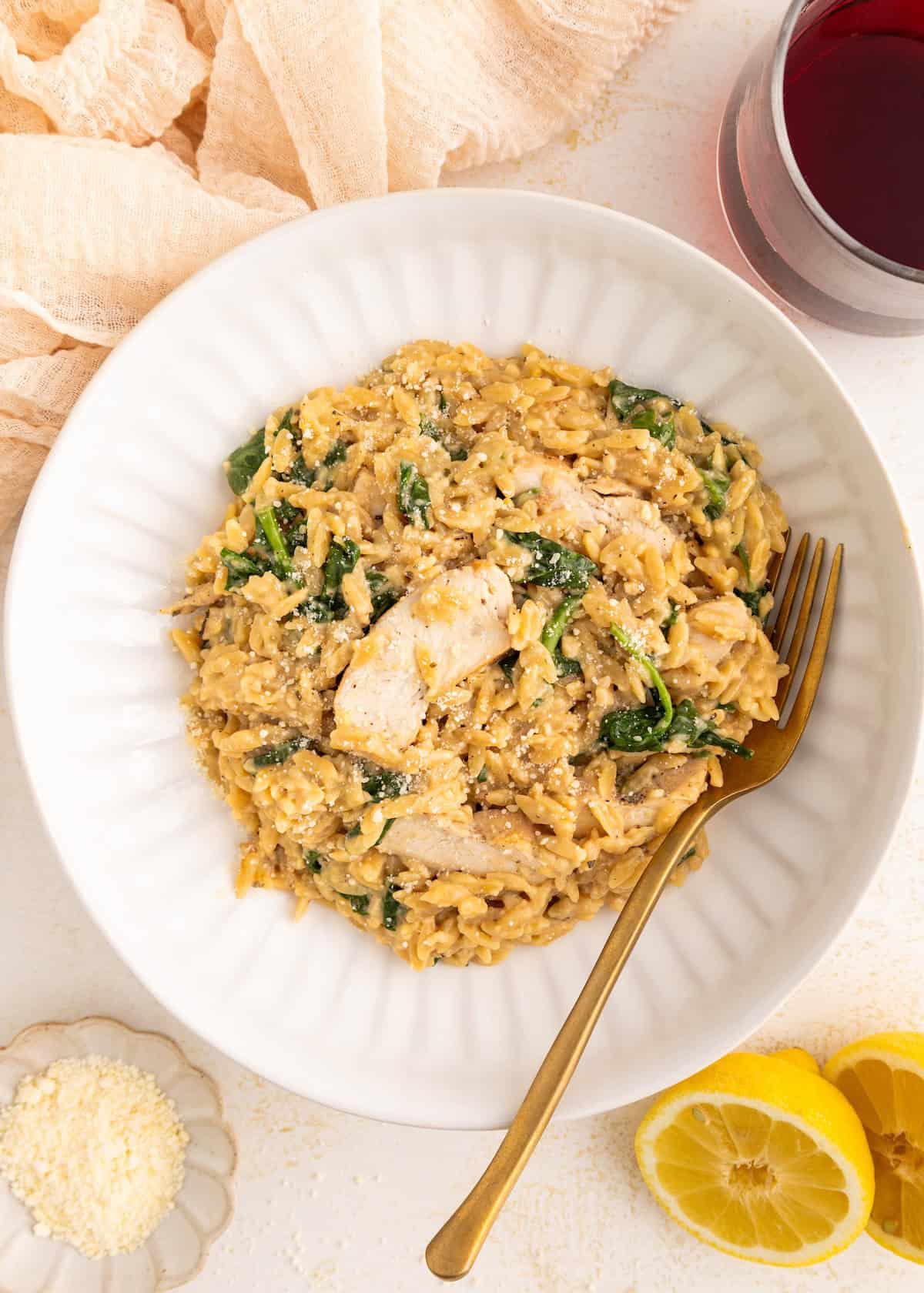 A bowl of creamy chicken and spinach orzo with a gold fork, next to lemon slices and red wine.