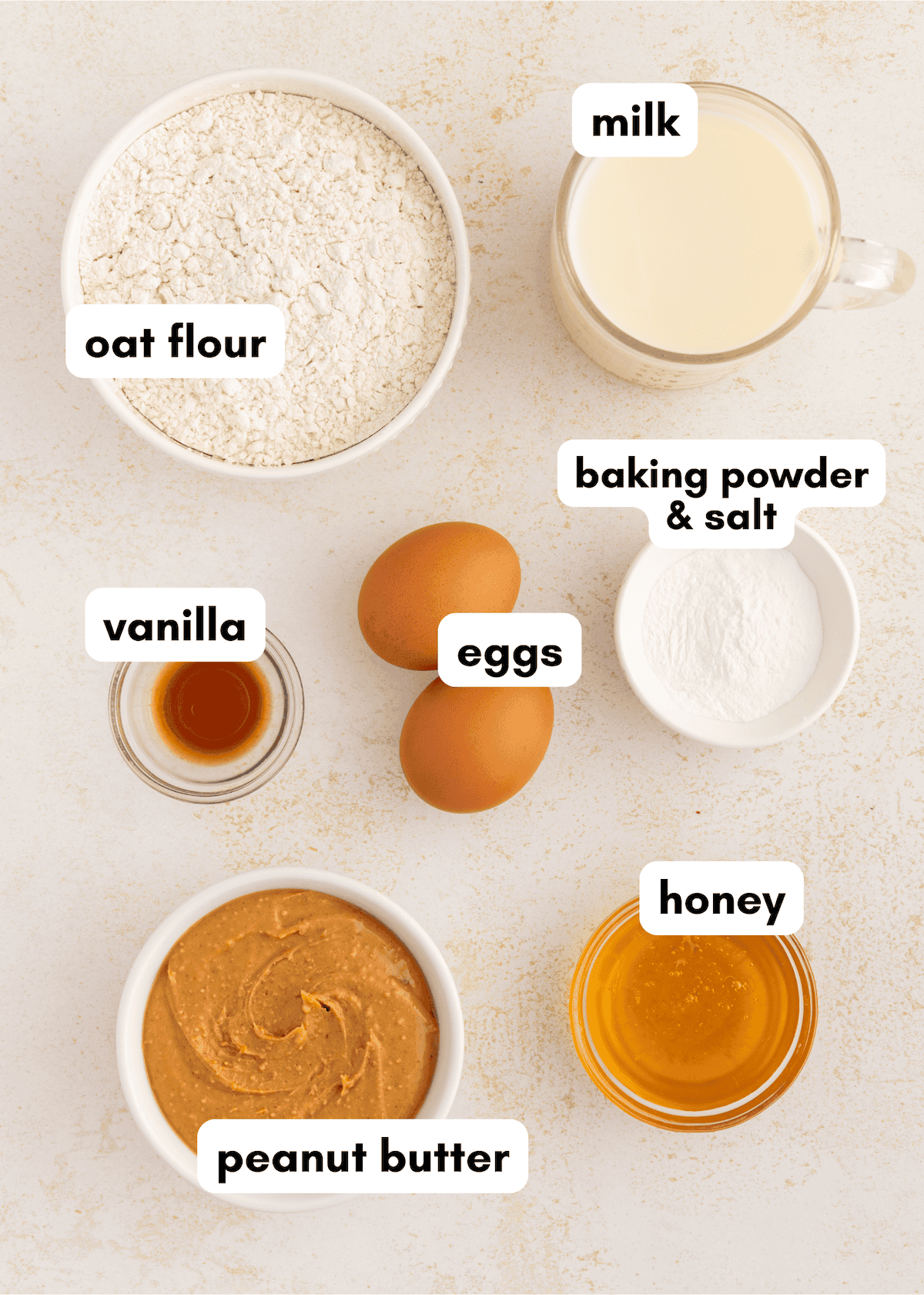 Top-down view of labeled baking ingredients for peanut butter waffles: oat flour, milk, eggs, vanilla, honey.