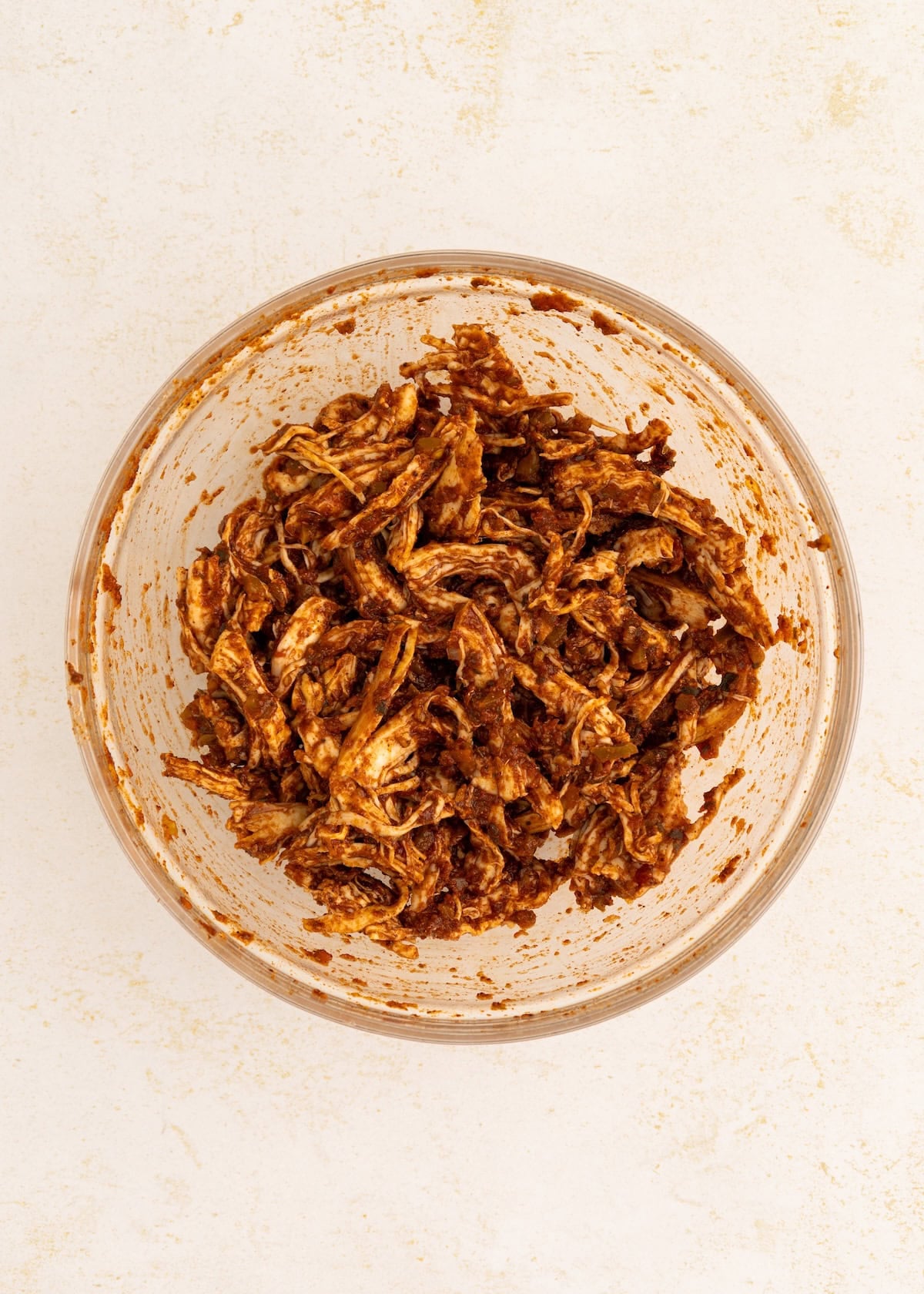 Shredded chicken coated in a dark red sauce in a glass bowl on a light surface.