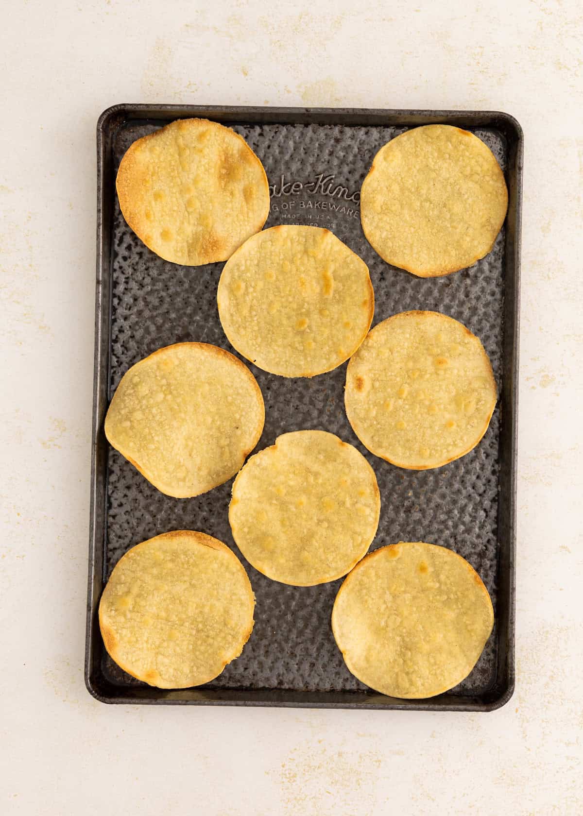 Eight baked corn tortillas on a dark baking sheet, arranged in rows.