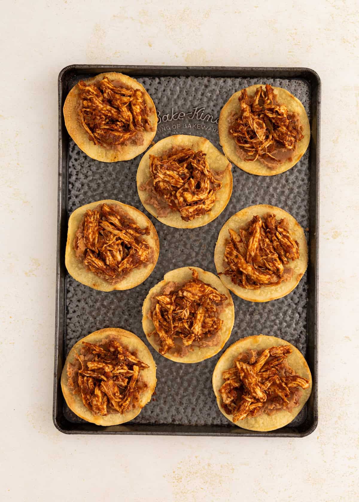 Nine small tostadas topped with shredded chicken are arranged on a baking sheet.