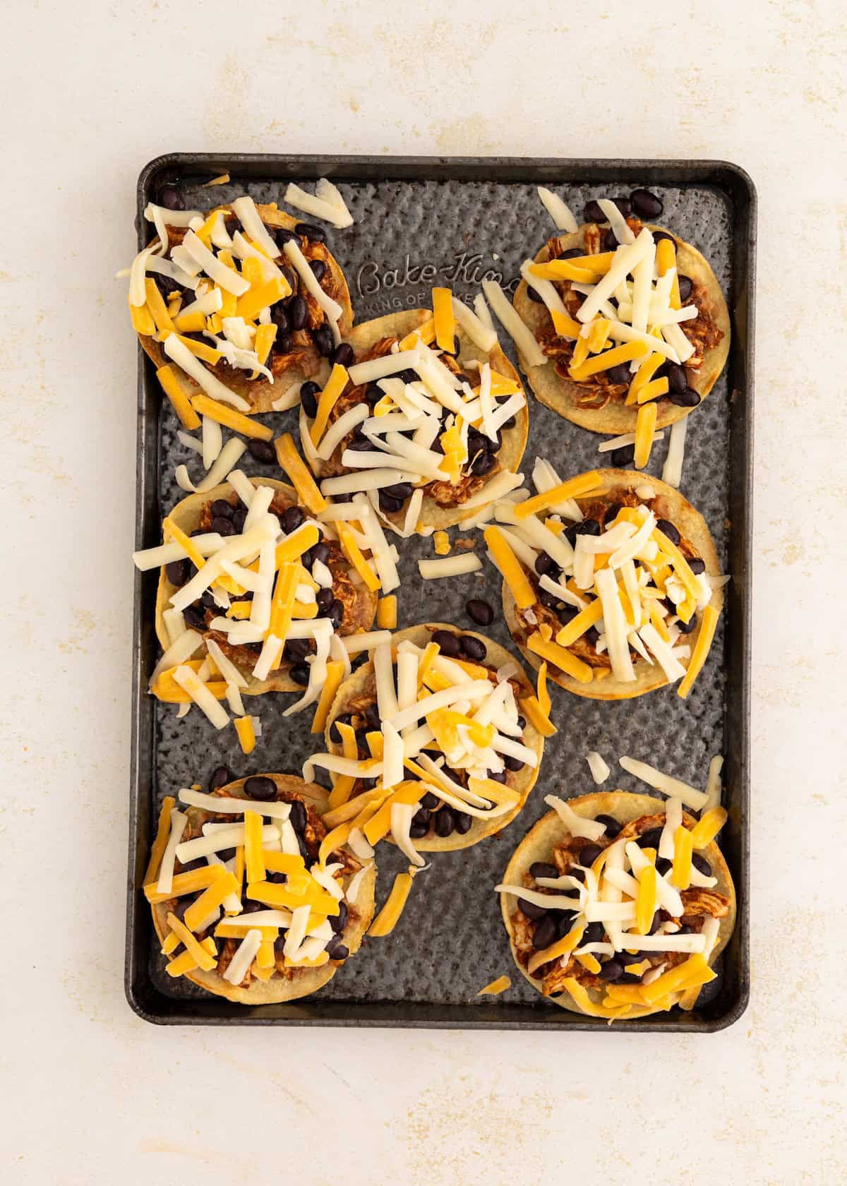 Mini tostadas with shredded cheese, black beans, and chicken on a baking sheet.