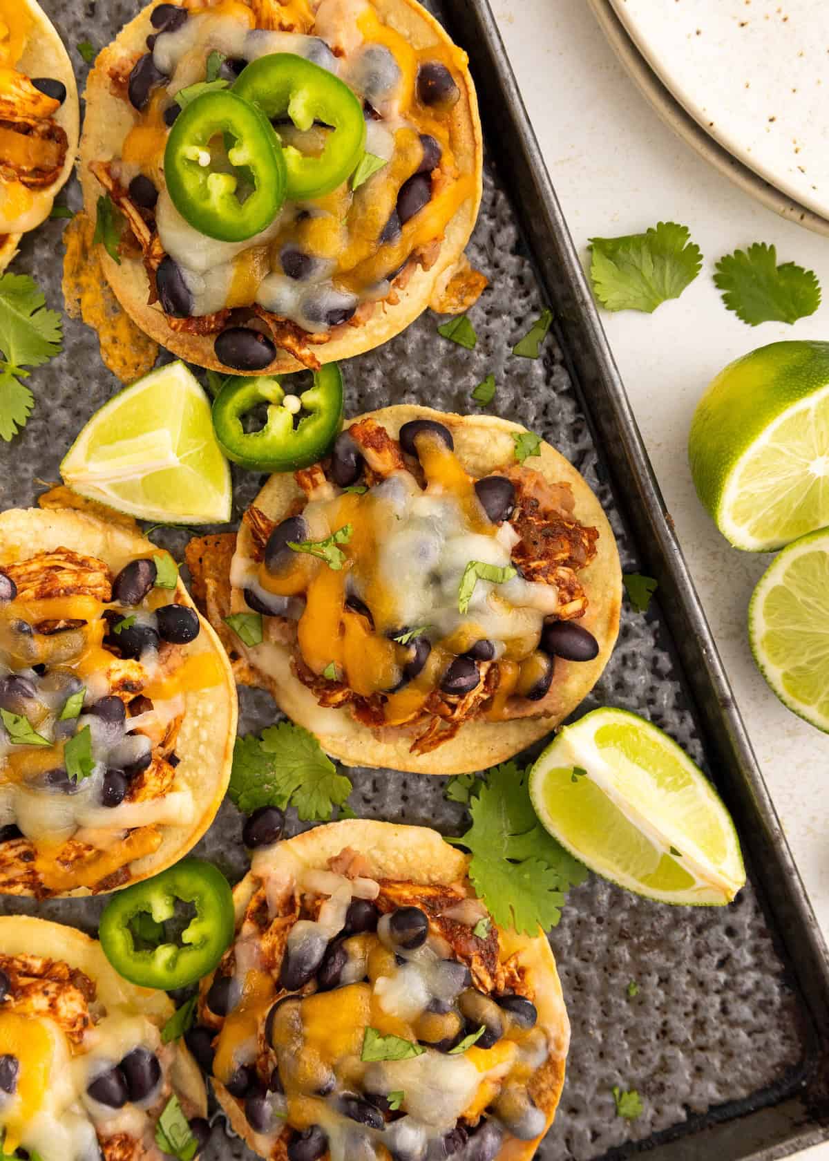 Mini tostadas with melted cheese, black beans, jalapeños, lime wedges, and cilantro on a tray.