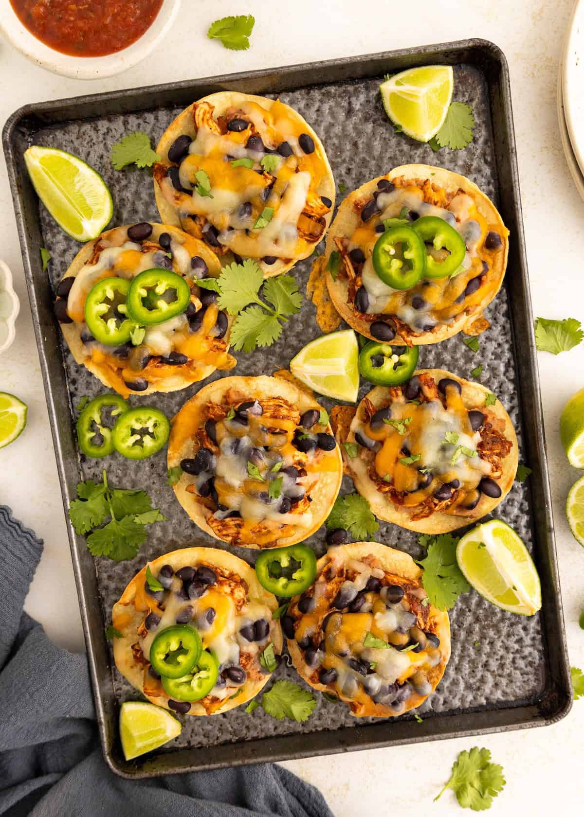 Mini pizzas with cheese, black beans, jalapeños, and lime wedges on a baking tray.