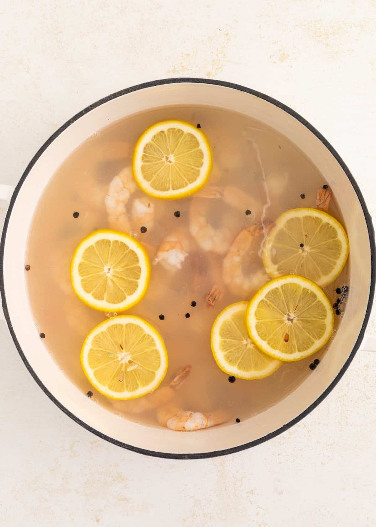 Shrimp boiling in a pot of water with lemon slices and black peppercorns.