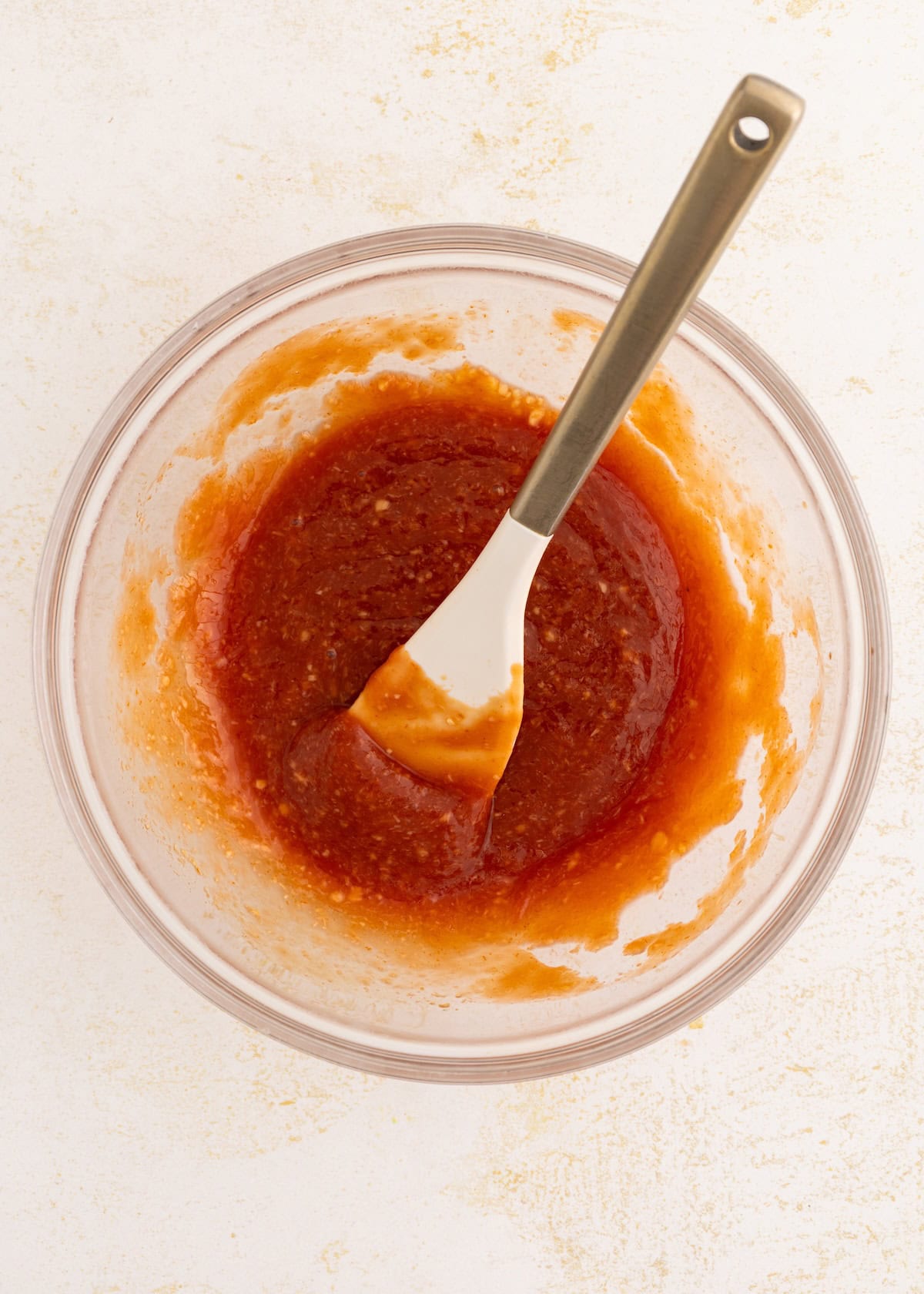 Glass bowl with red-orange sauce being mixed with a white spatula on a light surface.