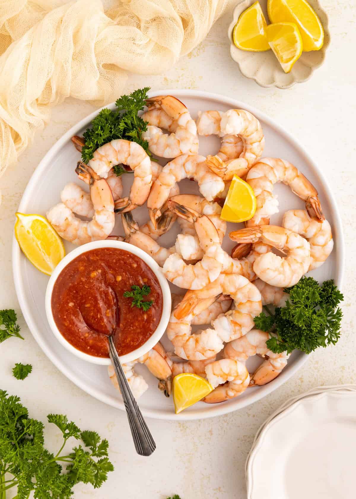 White plate of shrimp with cocktail sauce, lemon wedges, and parsley garnish on a table.