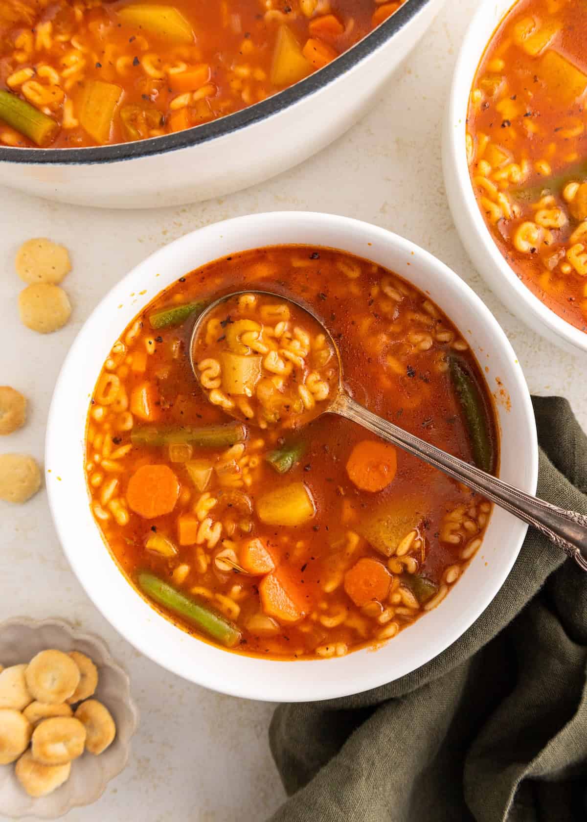 Bowl of vegetable soup with pasta and a spoon, next to oyster crackers on a light surface.