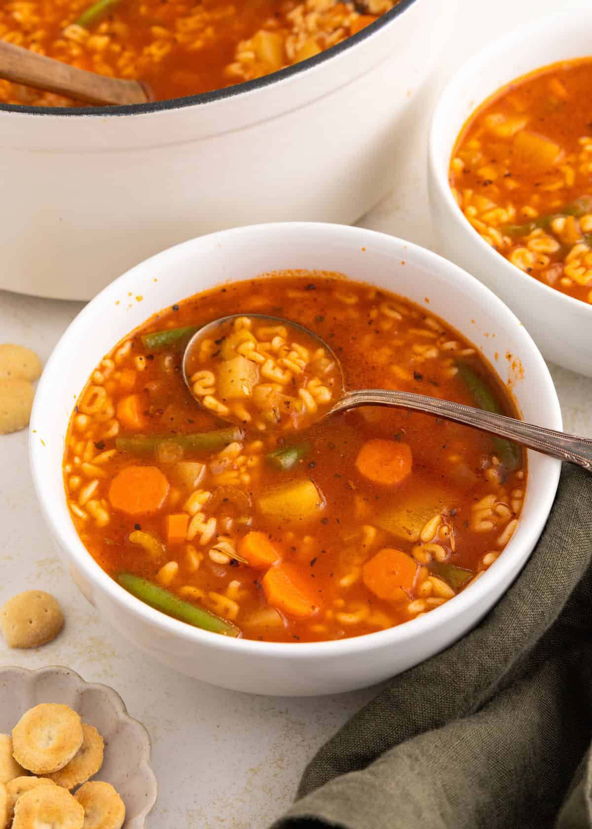 A bowl of vegetable soup with pasta and a spoon, surrounded by oyster crackers.