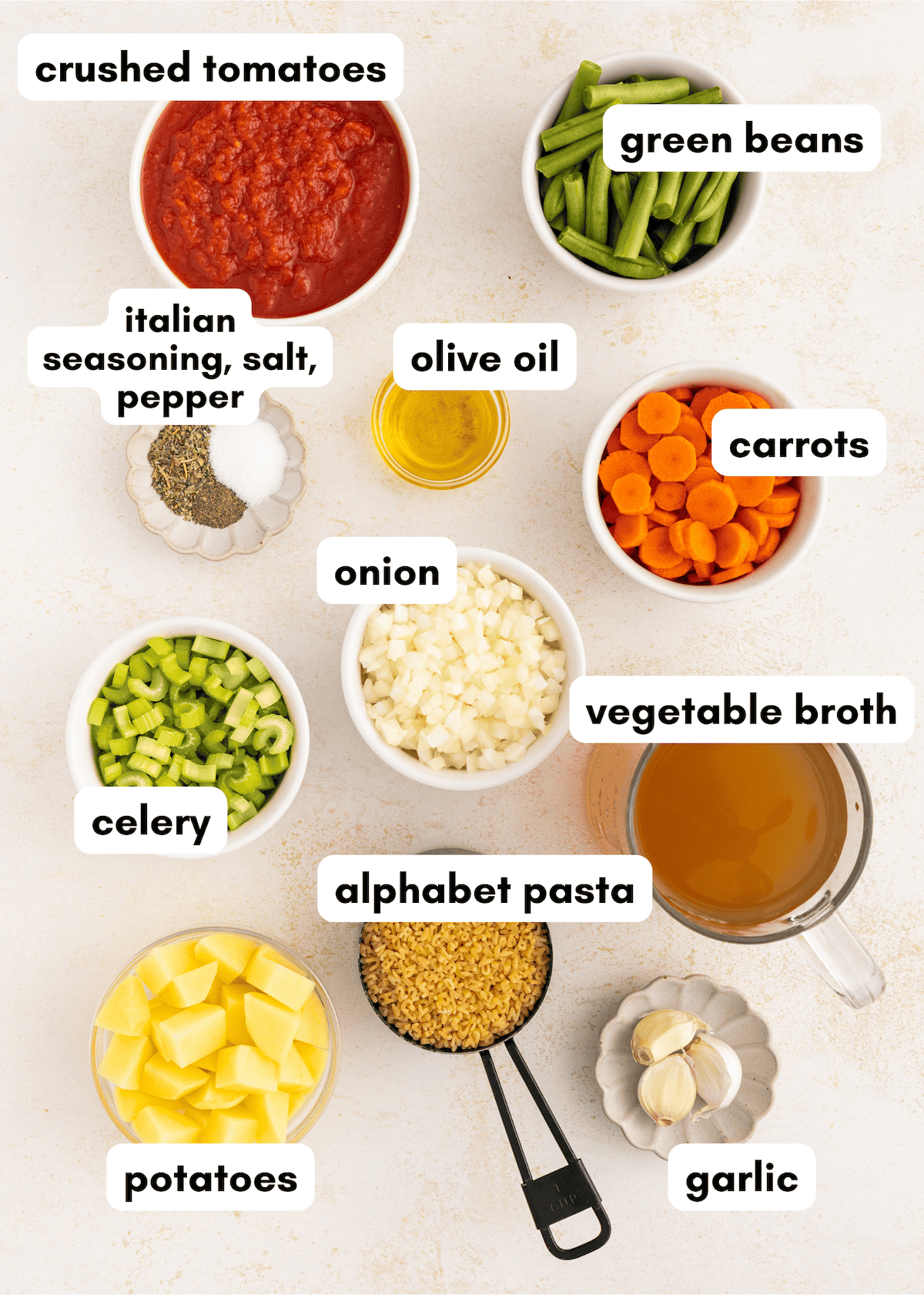 Bowls of chopped vegetables, alphabet pasta, broth, and seasonings arranged on a white surface.