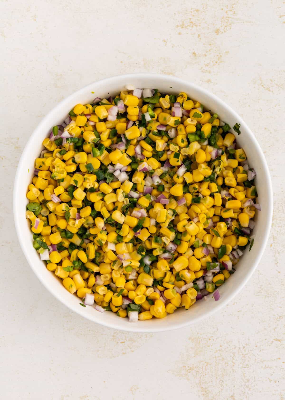 A bowl of corn salad with chopped red onion and herbs on a light background.