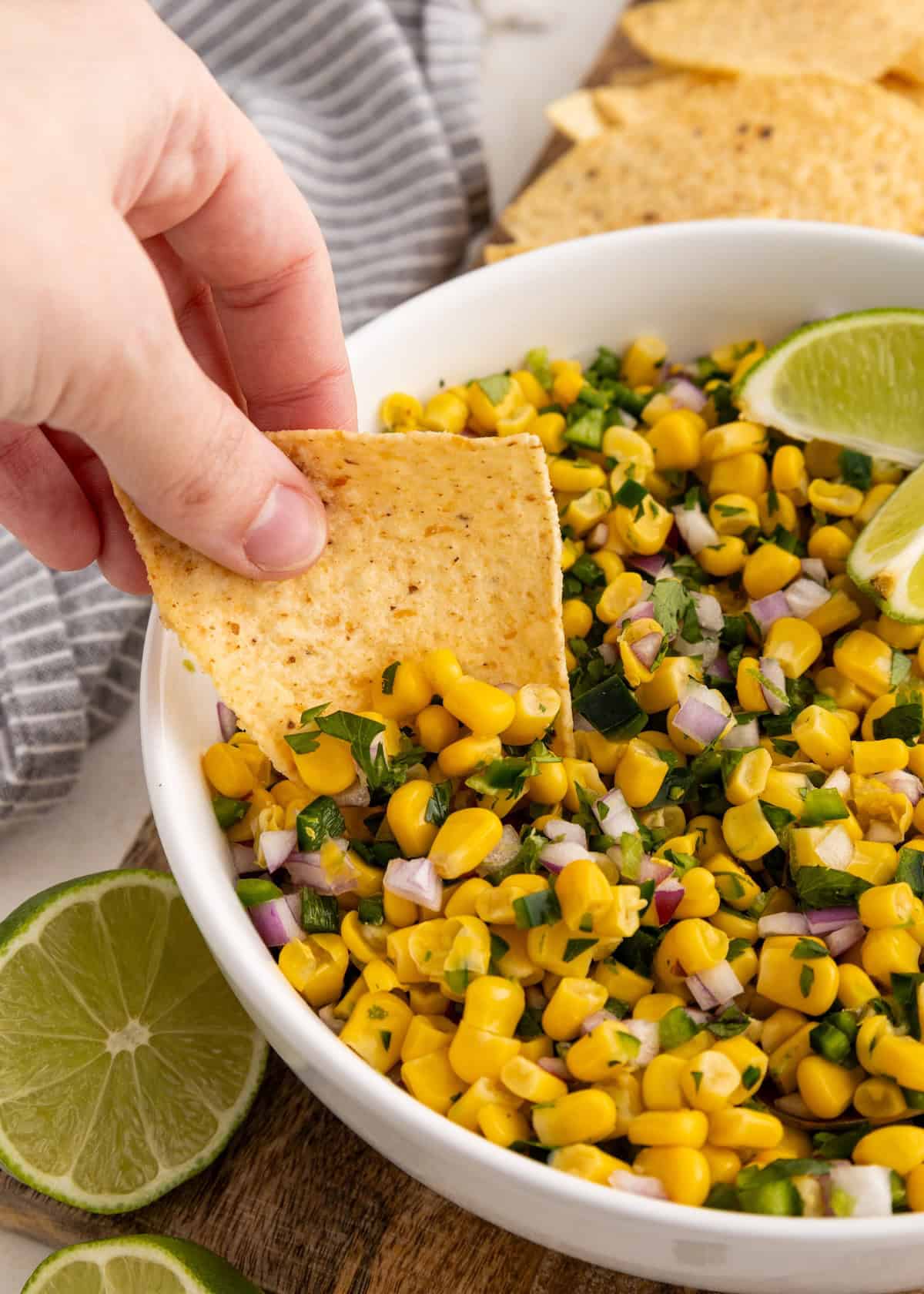 Hand dipping a tortilla chip into a bowl of corn salsa with lime wedges on the side.