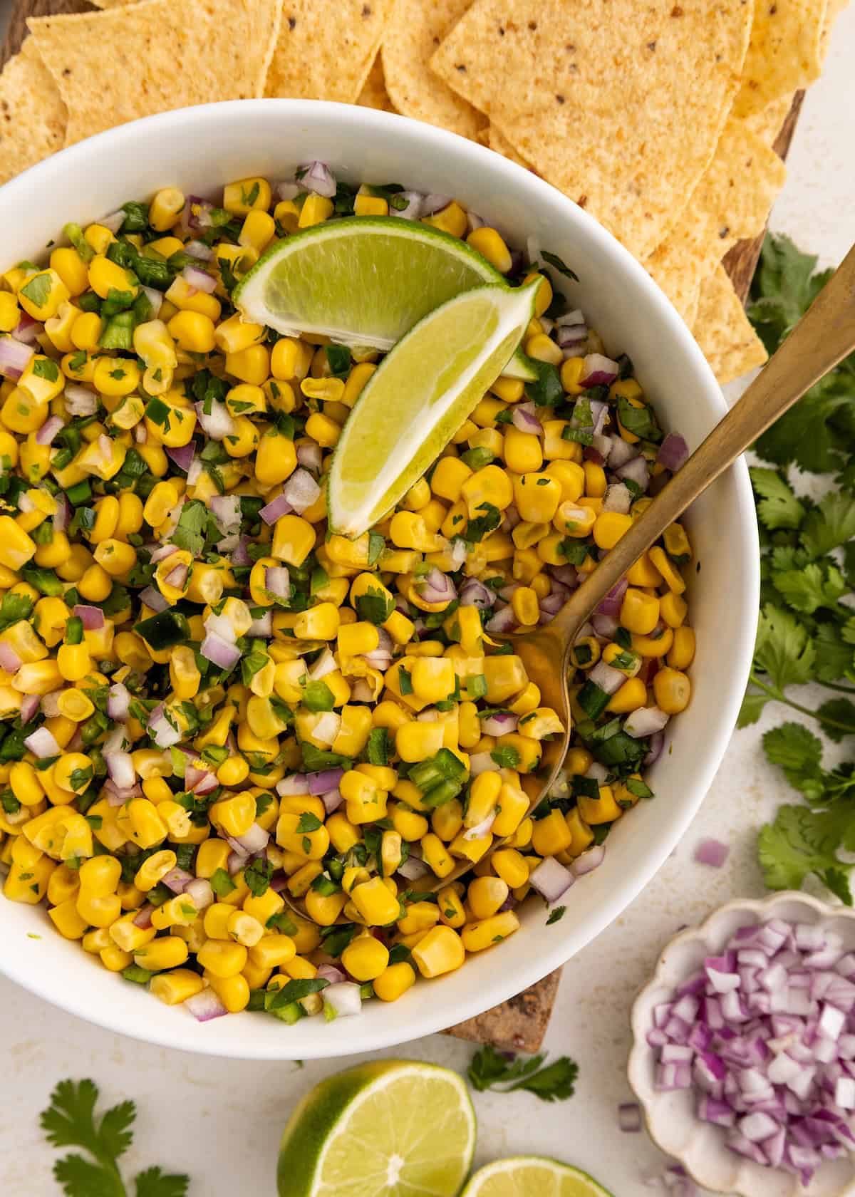 Bowl of corn salsa with lime wedges, tortilla chips, and fresh cilantro nearby.