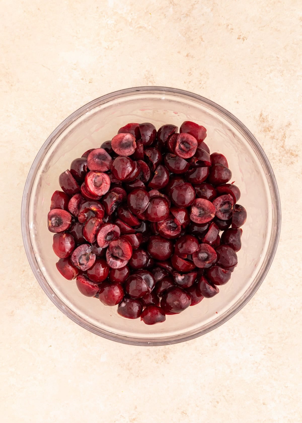 A glass bowl filled with halved, pitted cherries on a light-colored surface.